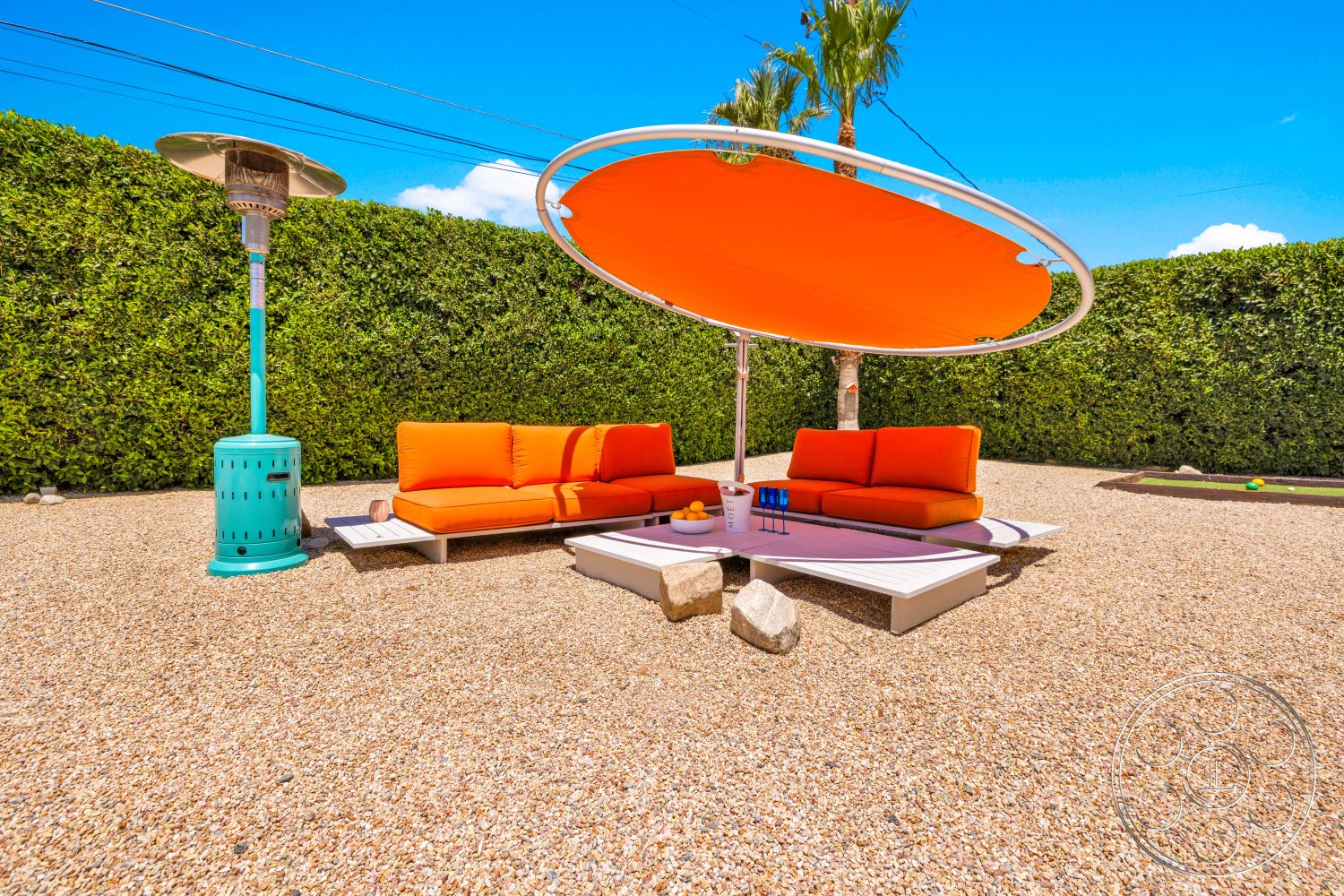 Palm Springs 76 - exterior, metal outdoor heater, contemporary outdoor lounge area, sunny backyard setting, green hedge privacy screen, orange outdoor sofa, tan gravel patio flooring, modern umbrella shade