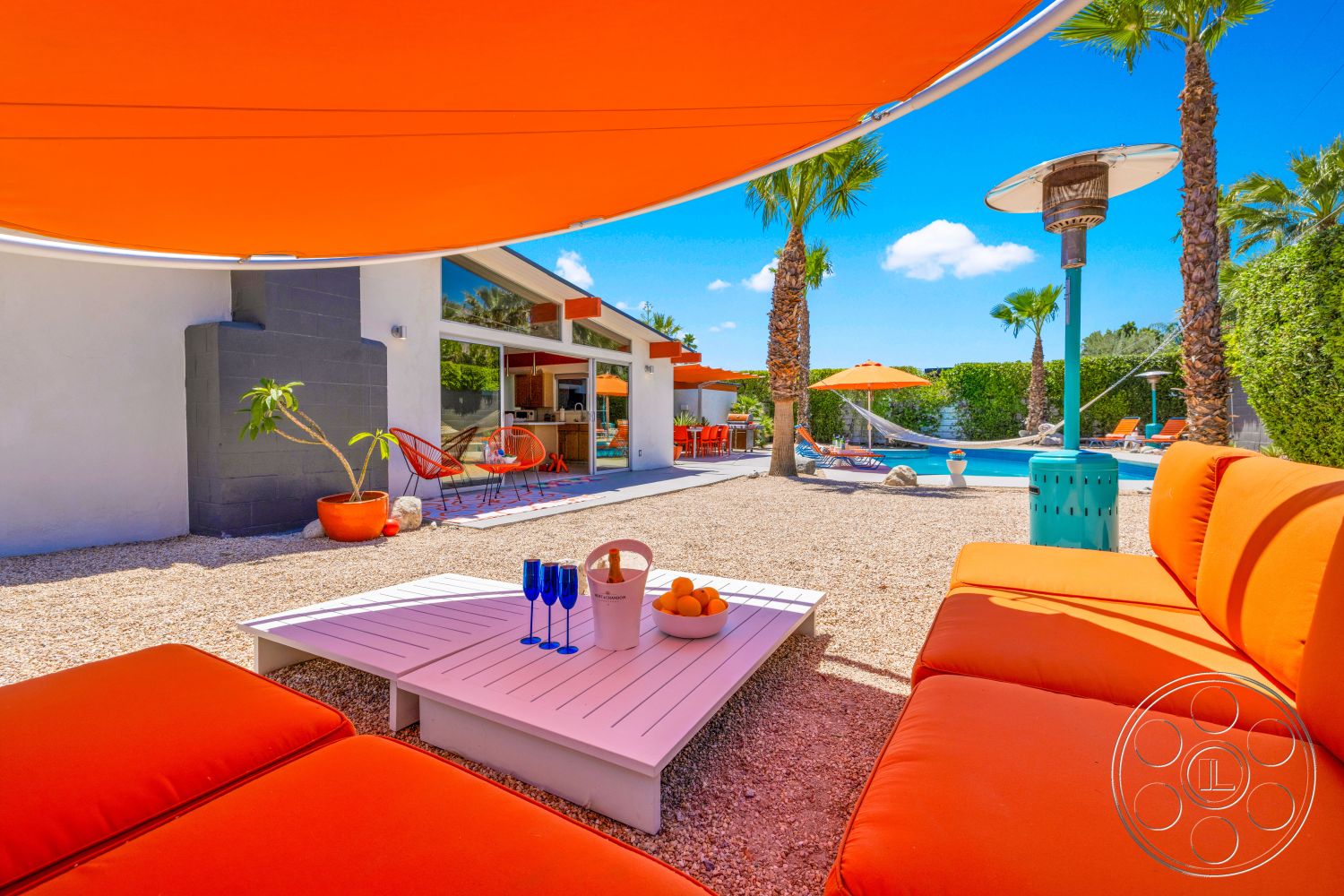 Palm Springs 76 - exterior, glass sliding doors, contemporary architecture, modern lounge area, gravel landscape, outdoor heating lamp, concrete patio flooring, palm trees landscaping, white stucco exterior walls, umbrella shade structure, colorful outdoor dining set, fiberglass swimming pool, orange fabric upholstery, white wooden coffee table, hammock resting area