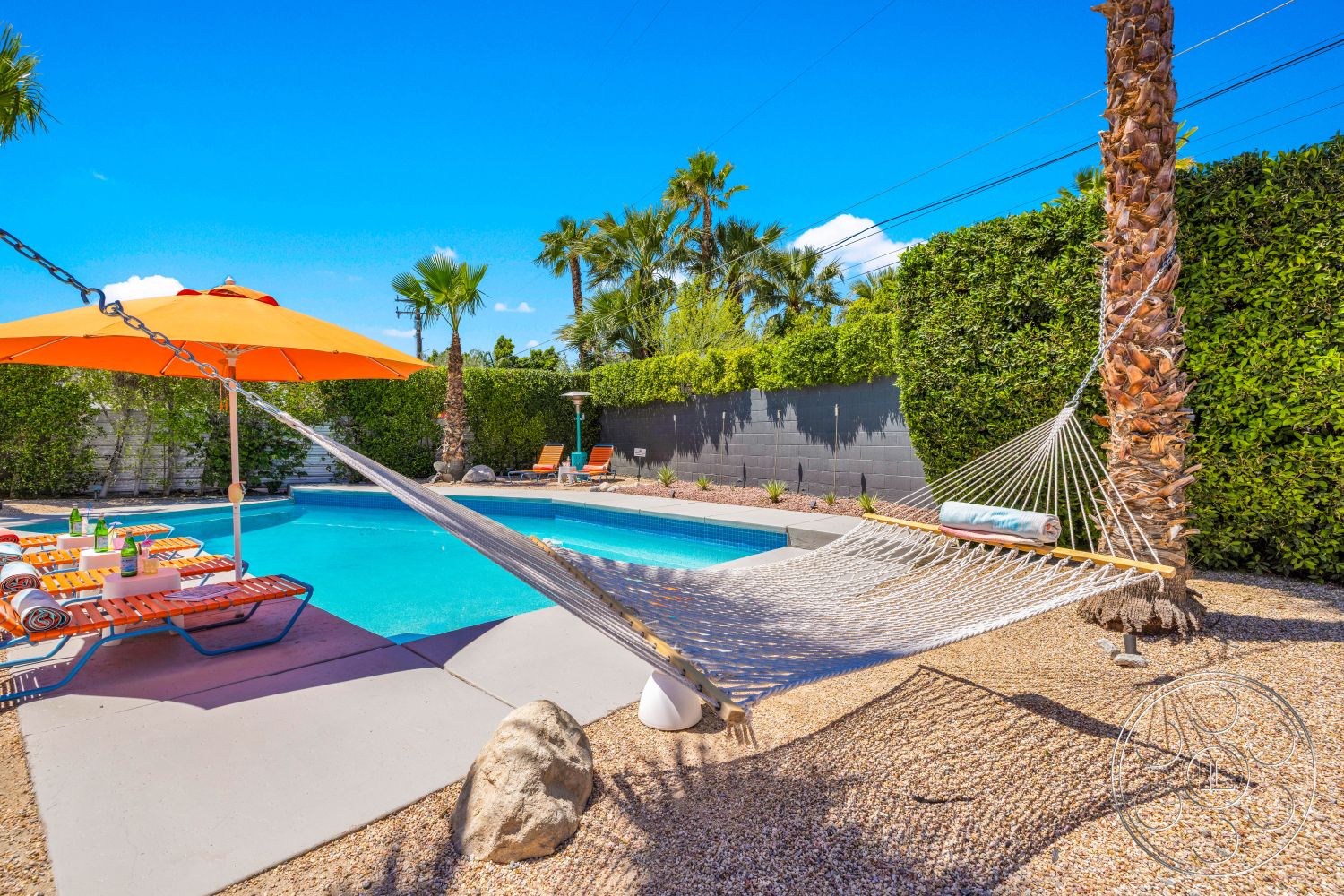 Palm Springs 76 - exterior, lounge chairs, gravel landscaping, concrete pool deck, low hedges, rectangular pool, outdoor dining table, sunny outdoor setting, concrete patio floor, palm tree landscaping, blue sky backdrop, orange patio umbrellas, hammock with rope details