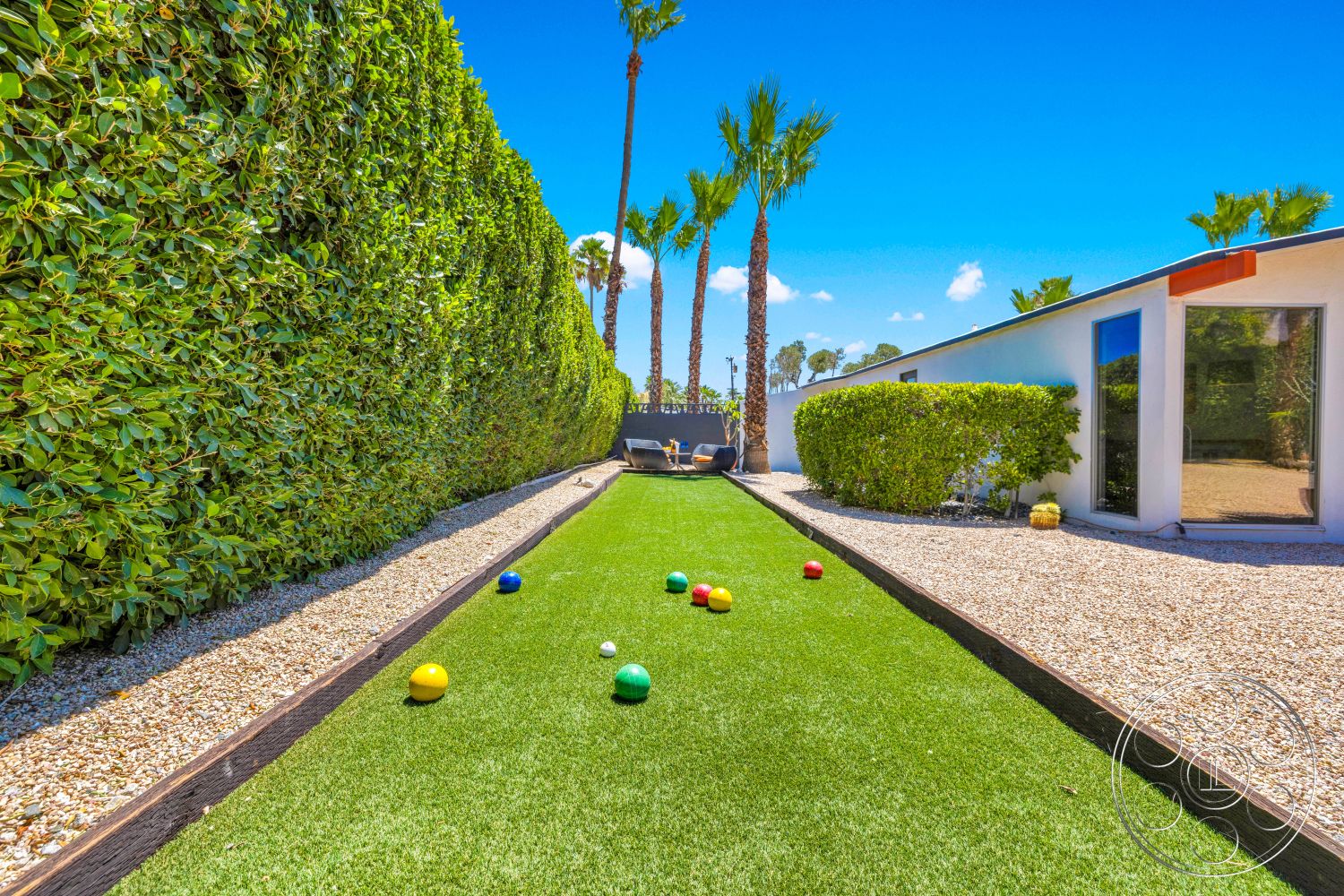 Palm Springs 76 - exterior, outdoor seating area, floor-to-ceiling windows, blue sky, brick edging, tropical palm trees, artificial turf lawn, modern home facade, decorative gravel pathway, sunny desert setting, hedge wall, lawn bowling game area