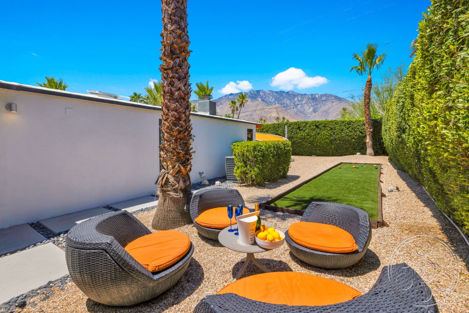 Palm Springs 76 - exterior, palm trees, circular seating arrangement, gravel landscaping, concrete patio floor, woven outdoor seating, desert mountain view, lush hedge privacy wall, orange cushion accents, green artificial turf lawn, colorful outdoor decor