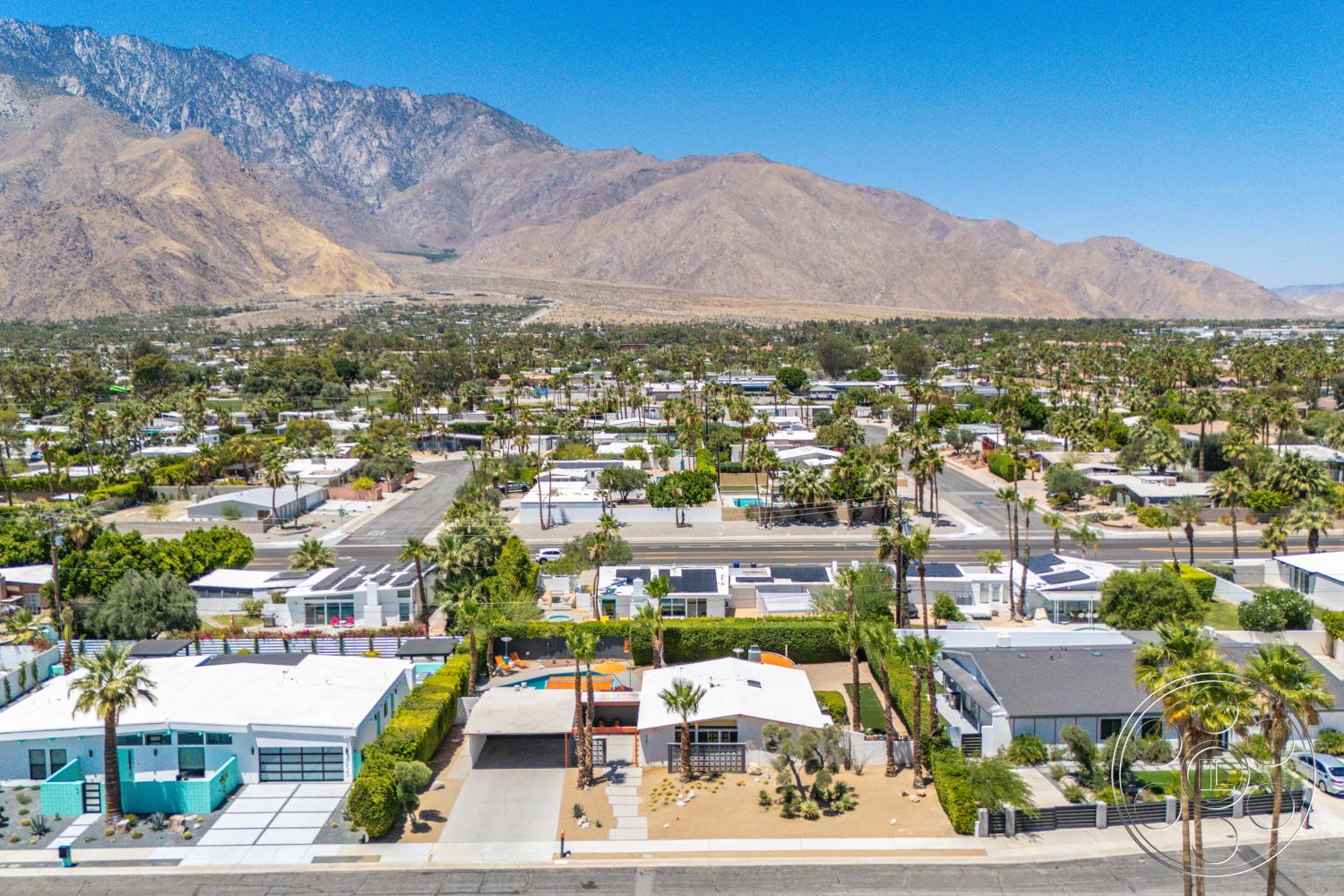 Palm Springs 76 - exterior, white stucco walls, concrete pathways, mountain backdrop, urban residential area, circular pool, landscaped garden areas, single-story homes, desert landscaping with palm trees, flat roof modern homes, concrete driveways, paved driveway with geometric pattern, open garage spaces, solar panel rooftops, green lawn areas, patio areas with outdoor seating