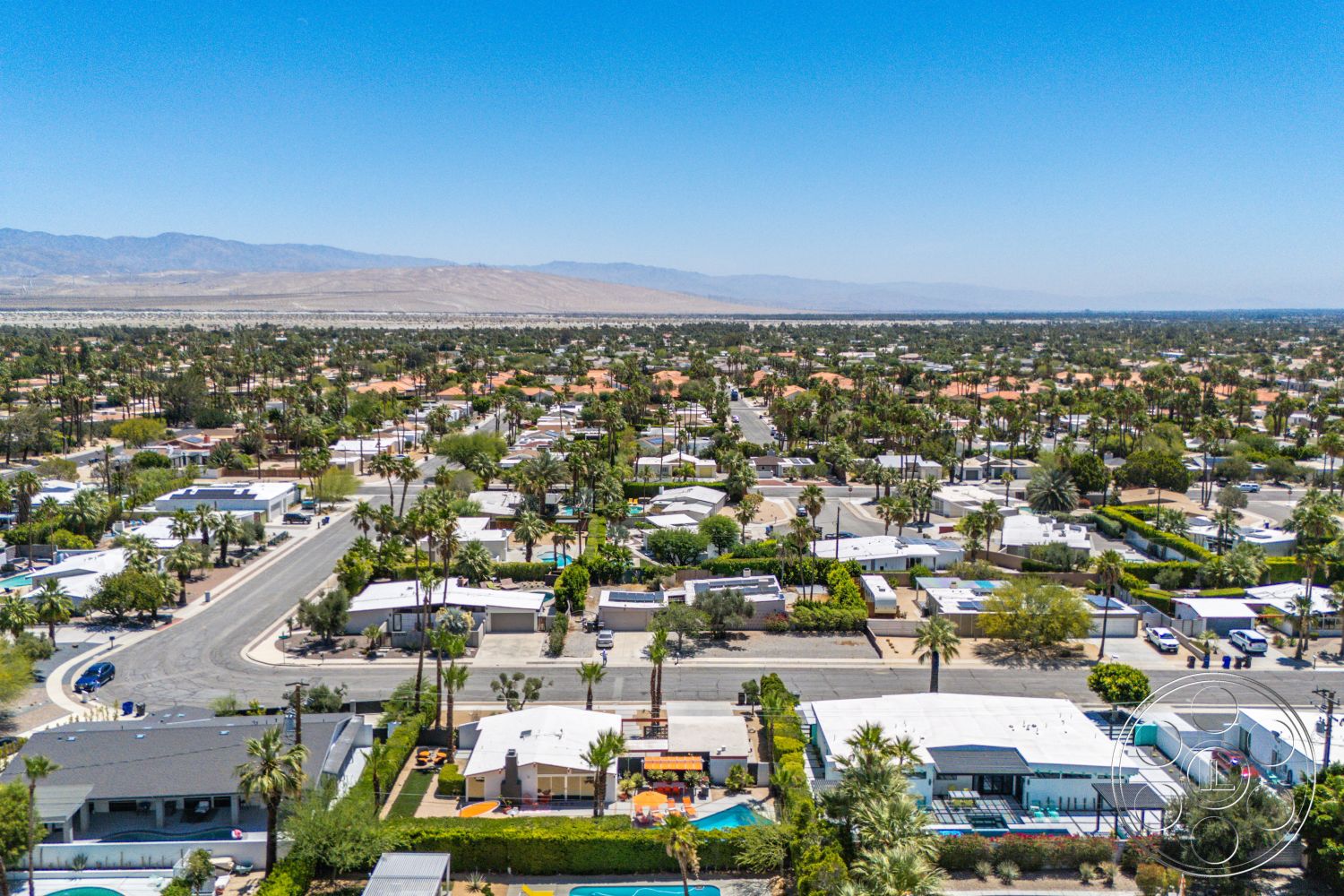 Palm Springs 76 - green vegetation, bushes and shrubs, mountain backdrop, blue sky, desert landscape, urban residential setting, palm tree landscaping, concrete driveways, aerial view of suburban neighborhood, single-story ranch-style homes, tile roofs, swimming pools, outdoor patios, gray asphalt roads, interior neighborhood streets, minimal front yard spaces, split-level homes