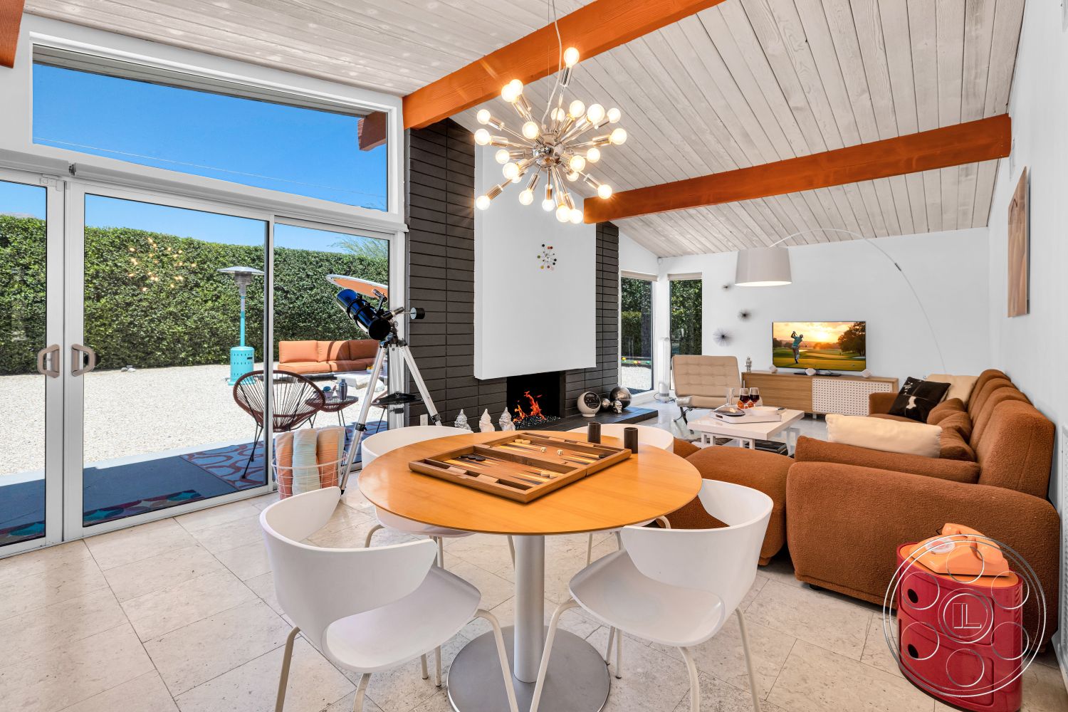 Palm Springs 76 - large glass sliding doors, textured accent wall, modern entertainment unit, rectangular dining table, light-colored tile flooring, wood beam ceiling, white painted walls, stone fireplace wall, potted indoor plants, contemporary light fixture, white plastic dining chairs, tan upholstered sectional sofa, various outdoor furniture visible through glass doors