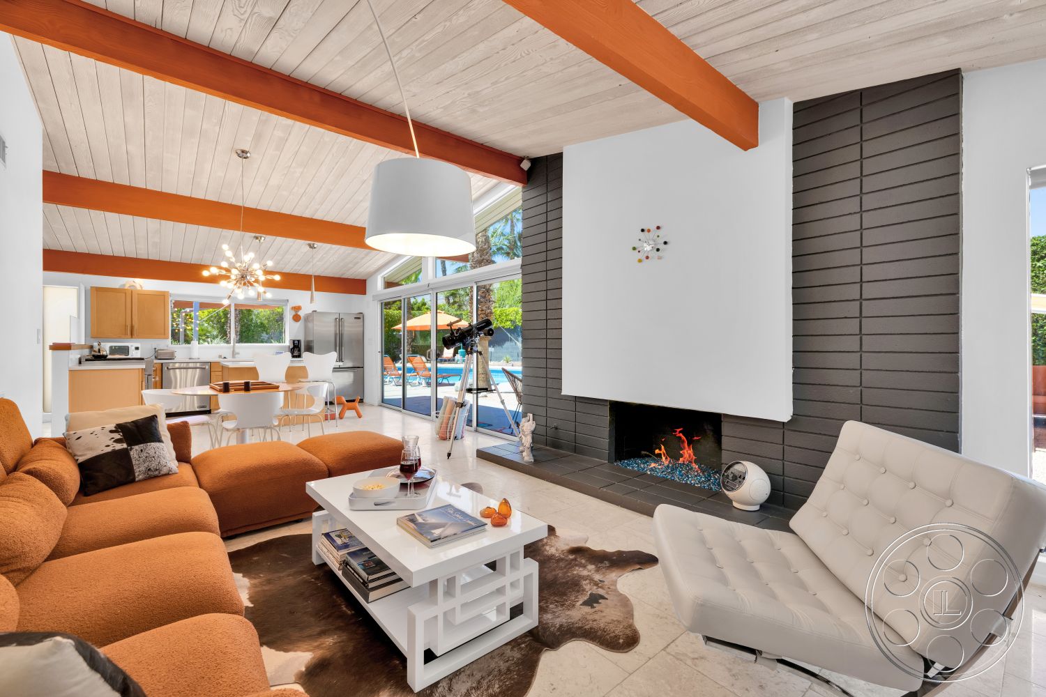 Palm Springs 76 - exterior, stainless steel appliances, glass sliding doors, rounded dining table, white dining chairs, tan sectional sofa, exposed wood ceiling beams, mid-century modern architectural style, open concept kitchen, wooden kitchen cabinets, wood beam ceiling, white painted walls, vaulted living room ceiling, palm tree landscaping, modern chandelier lighting, tile living room flooring, outdoor pool area view, modern white coffee table, concrete fireplace surround, black stone feature wall
