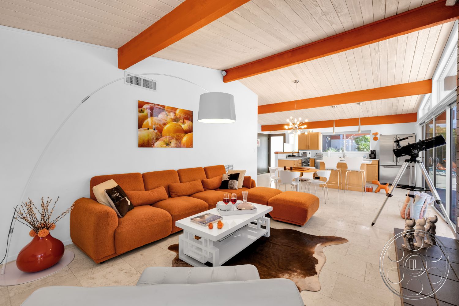 Palm Springs 76 - wood dining table, glass sliding doors, kitchen island, vaulted ceiling, cream-colored walls, white dining chairs, light-colored tile flooring, white coffee table, exposed wood ceiling beams, modern pendant light fixture, orange fabric sectional sofa, orange-painted beams, decorative cowhide rug, appliances in kitchen area