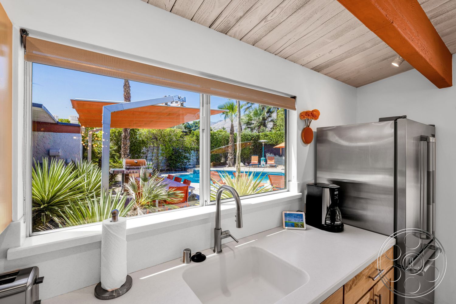 Palm Springs 76 - wood cabinetry, bright natural lighting, stainless steel refrigerator, kitchen sink, exposed wood ceiling beams, contemporary kitchen design, large picture window, white painted walls, white quartz countertops, suburban backyard view