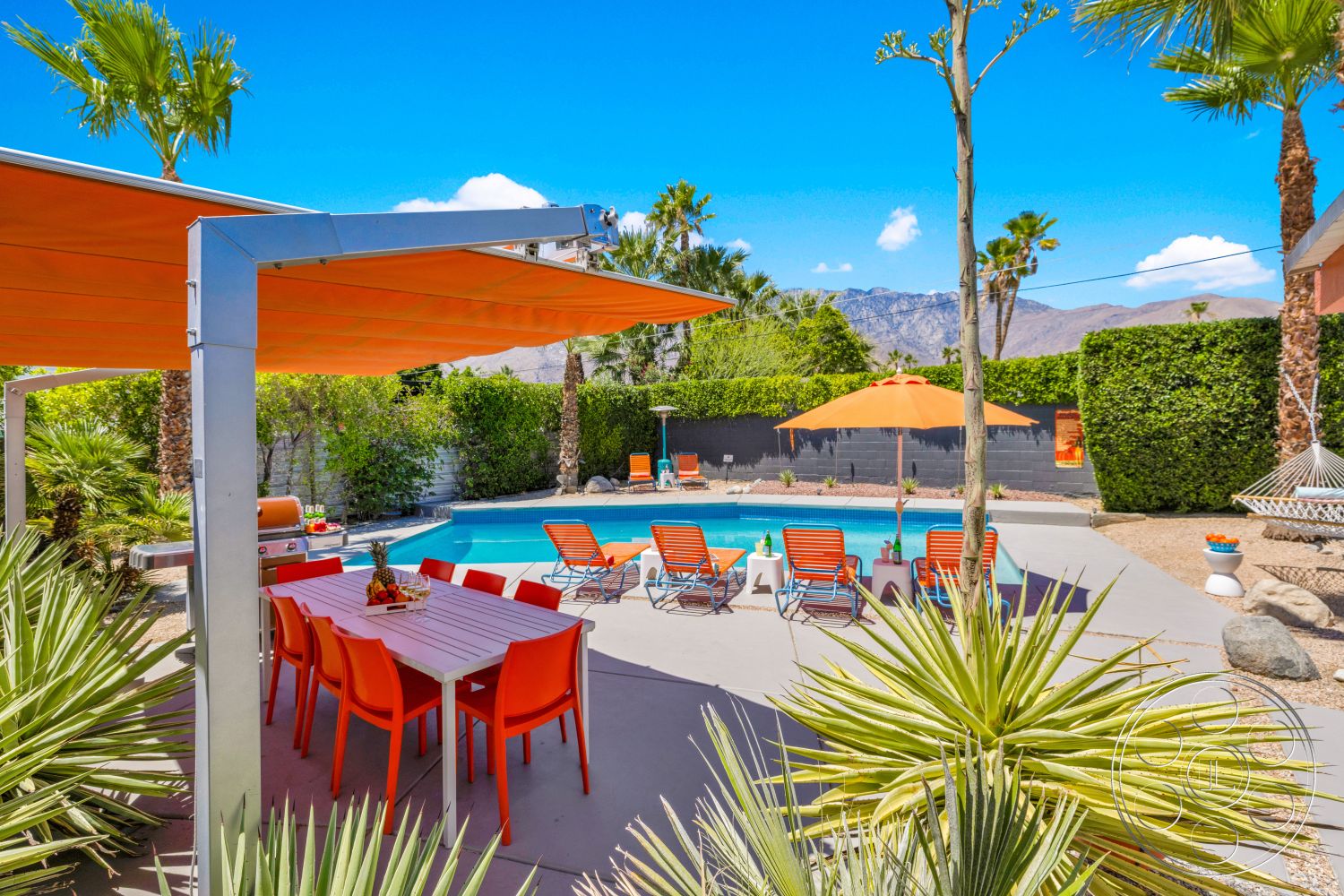 Palm Springs 76 - exterior, palm trees, mountain backdrop, rectangular pool, modern outdoor grill, landscaped garden area, concrete patio flooring, steel patio furniture, textured garden wall, sunny desert setting, orange plastic dining chairs, bright orange patio umbrellas, gray stone pool border, cement outdoor dining table