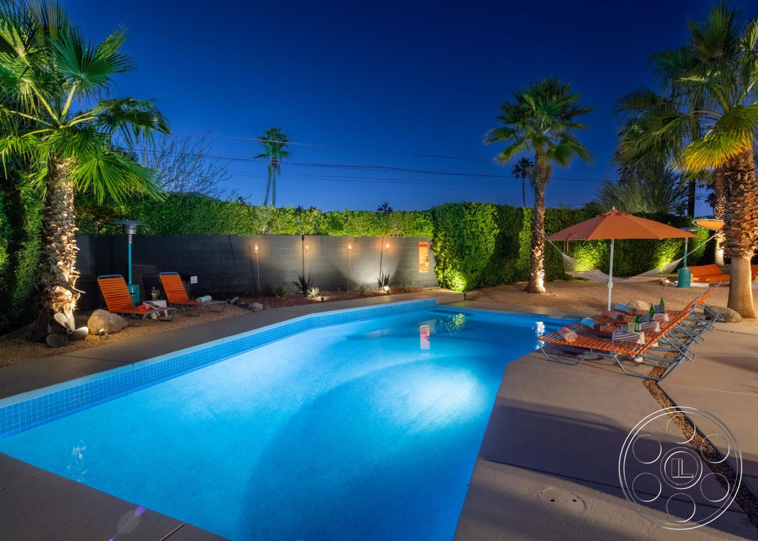 Palm Springs 76 - exterior, palm trees, landscaped yard, lush green hedges, rectangular pool, outdoor lighting fixtures, tiled pool edge, concrete pool border, concrete patio floor, orange patio umbrella, decorative gravel ground cover, multi-colored lounge chairs