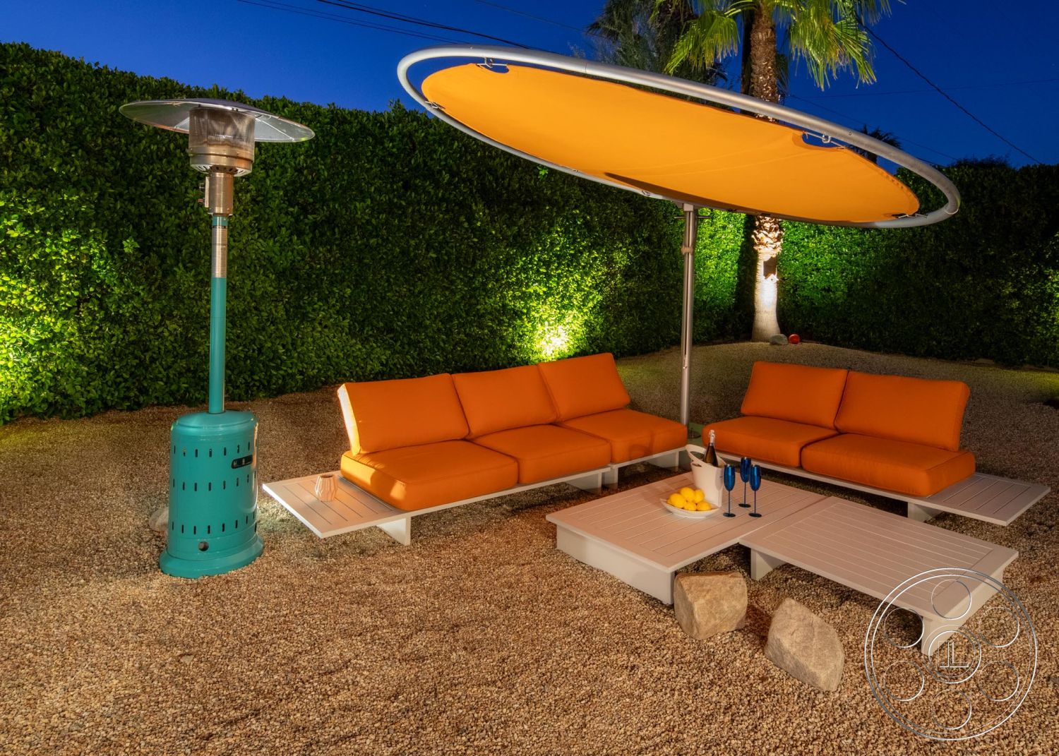 Palm Springs 76 - ambient lighting, outdoor lounge area, tropical palm trees, metal patio heater, gravel patio flooring, orange cushioned sectional sofa, white slatted coffee table, rounded fabric sunshade, landscaped green hedge backdrop