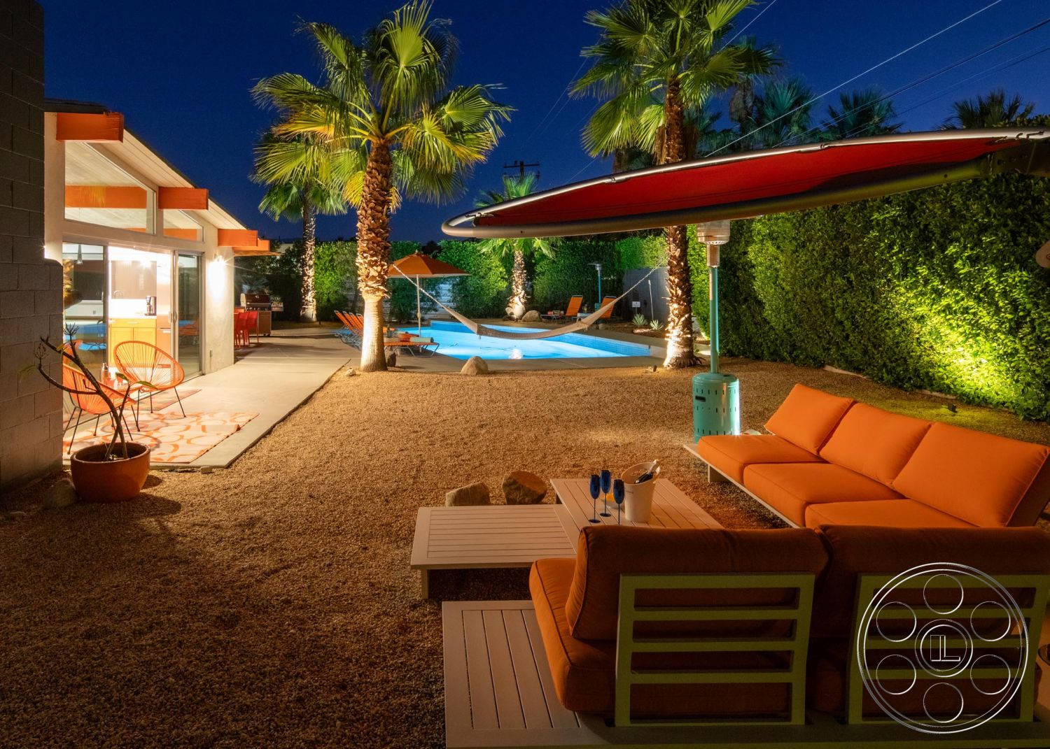 Palm Springs 76 - exterior, landscaped garden, palm trees, glass sliding doors, outdoor dining area, modern architectural style, rectangular pool, wooden patio furniture, cinder block wall, gravel patio flooring, orange patio umbrella, orange outdoor cushions, concrete pool surround, stone accent boulders, linear outdoor lighting