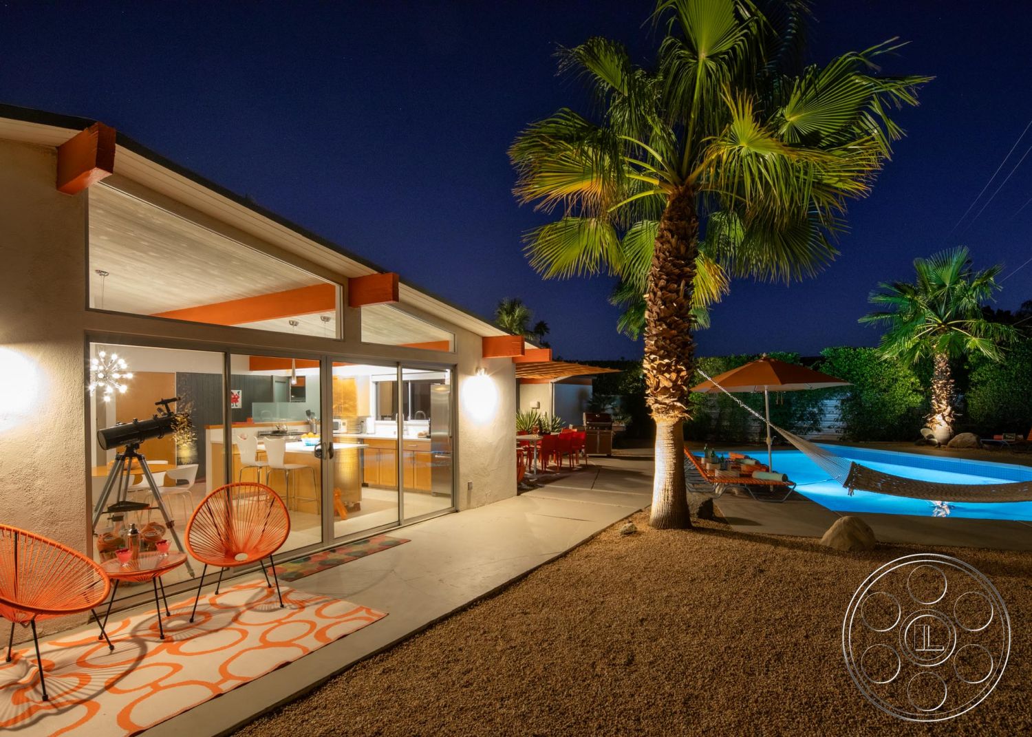 Palm Springs 76 - exterior, landscaped garden, palm trees, sliding glass doors, rectangular swimming pool, outdoor dining area, nighttime setting, modern architectural style, gravel landscaping, hammock, decorative outdoor rug, exposed wood ceiling beams, concrete patio floor, orange outdoor furniture