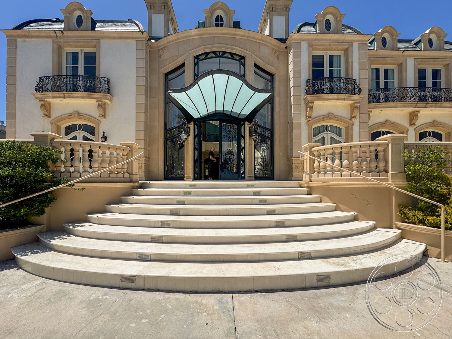 Tiffany Estate - exterior, symmetrical facade, arched doorways, landscaped shrubbery, decorative wrought iron railings, stone staircase with rounded steps, glass entryway awning, beige stucco facade with ornate detailing, sloped roof with dormer windows, balconies with wrought iron accents, elevated entrance platform