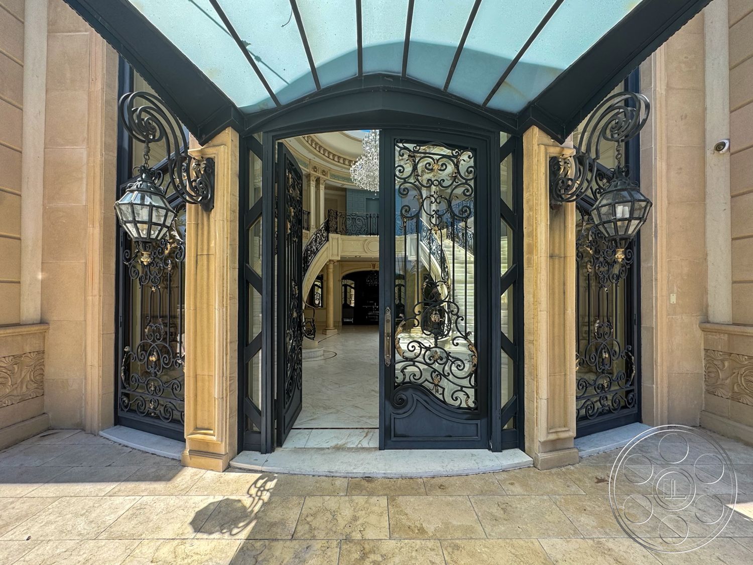 Tiffany Estate - exterior, decorative stone accents, chandelier lighting fixture, ornate wrought iron doors, tiled entryway flooring, cream stone walls, glass canopy roof, curved entry steps