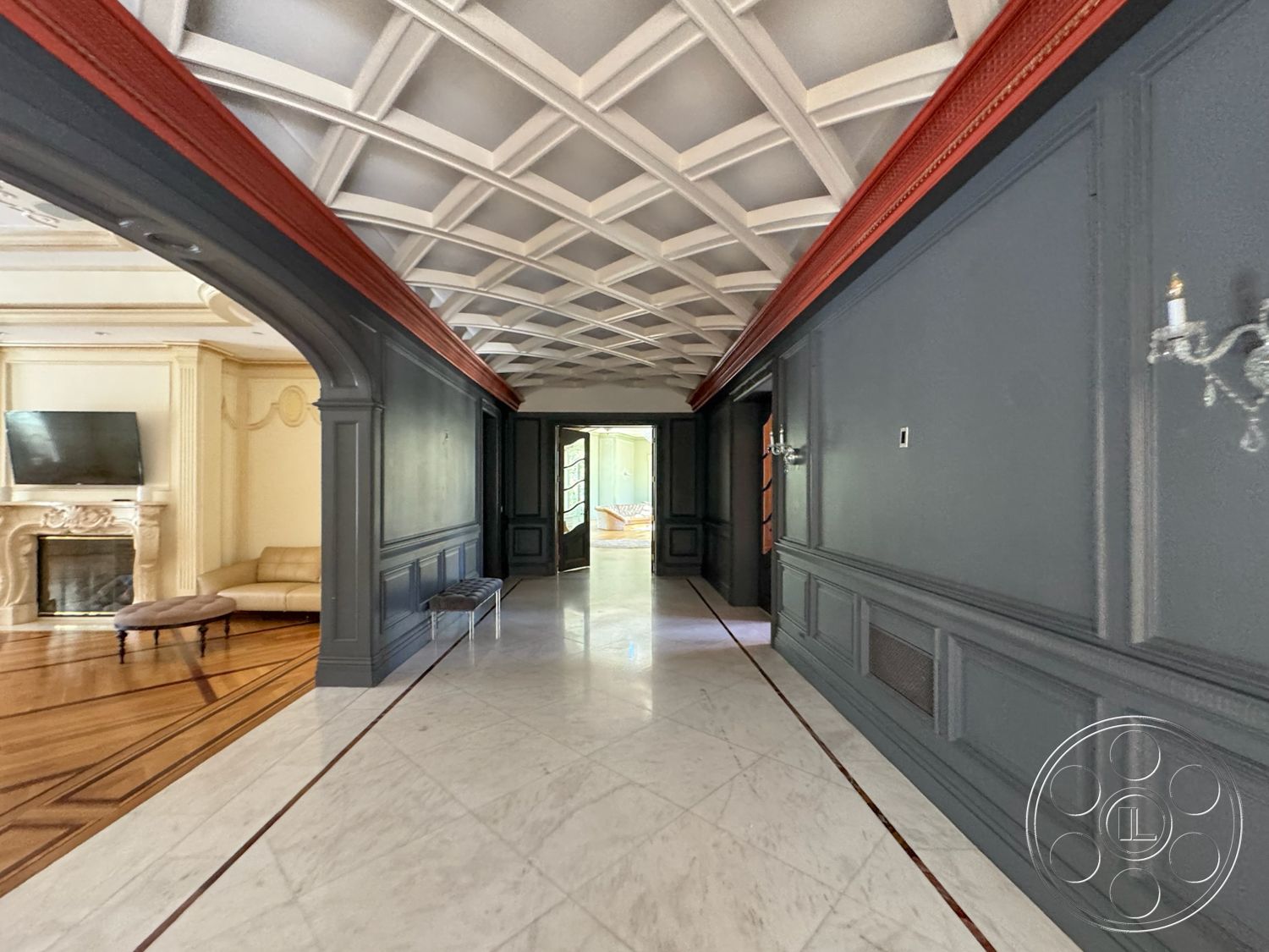 Tiffany Estate - arched doorways, dark wood paneling walls, decorative wall moldings, chevron patterned marble flooring, coffered ceiling with white and red detailing, ornate fireplace with cream stone surround, luxurious living room area, plush cream-colored sofa, floor-to-ceiling windows leading to outdoor space, light-filled hallway, intricate ceiling architecture