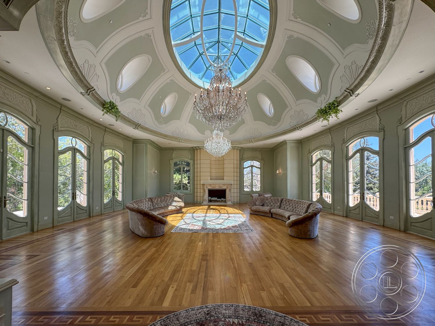 Tiffany Estate - light green painted walls, symmetrical room layout, vaulted ceiling with dome skylight, chandelier with crystal detailing, hardwood flooring with intricate inlay design, large arched windows with decorative framing, stone fireplace wall with smooth finish, soft, curved sofa seating, decorative wall molding with floral motifs, elegant area rug with intricate patterns