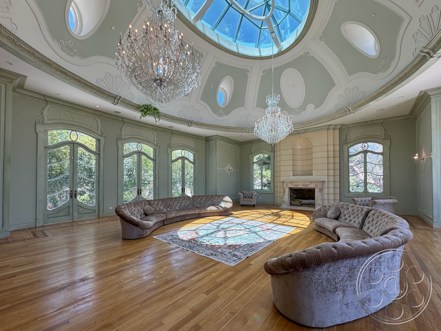 Tiffany Estate - hardwood flooring, stone fireplace, chandelier lighting, arched windows, decorative area rug with intricate pattern, vaulted ceiling with skylight, oval-shaped seating arrangement, detailed plaster ceiling moldings, beige velvet sofas, light green wall treatments, French doors leading to outdoor views