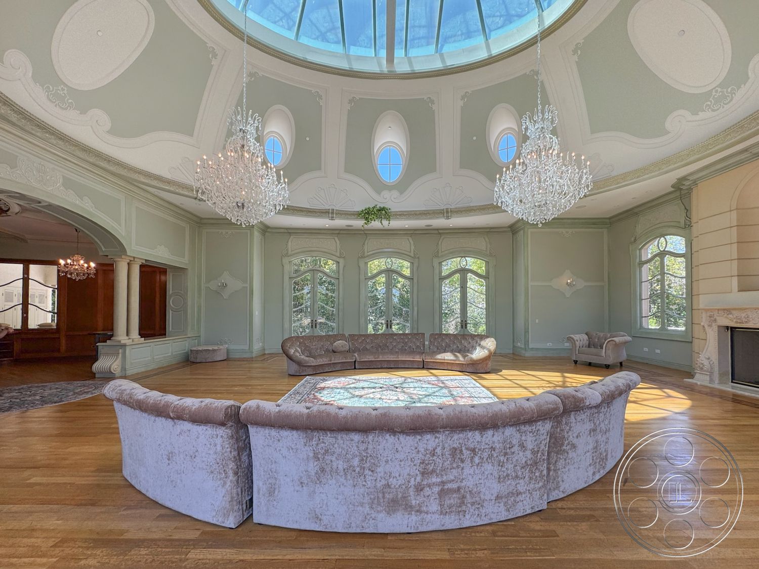 Tiffany Estate - natural light, hardwood flooring, stone fireplace, open floor plan, floor-to-ceiling windows, luxurious setting, elegant ambiance, round area rug, vaulted ceiling with skylight, chandelier lighting fixtures, ornate architectural details, plaster walls with decorative moldings, curved velvet sofa