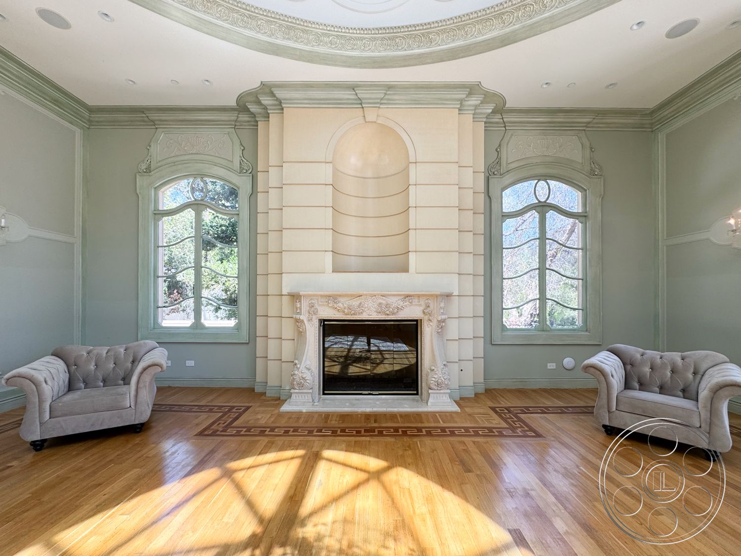 Tiffany Estate - hardwood flooring, textured wall panels, arched windows, white window trim, marble fireplace surround, light green painted walls, ornate ceiling crown molding, two tufted armchairs, decorative wood inlay floor design, elegant chandelier light fixture