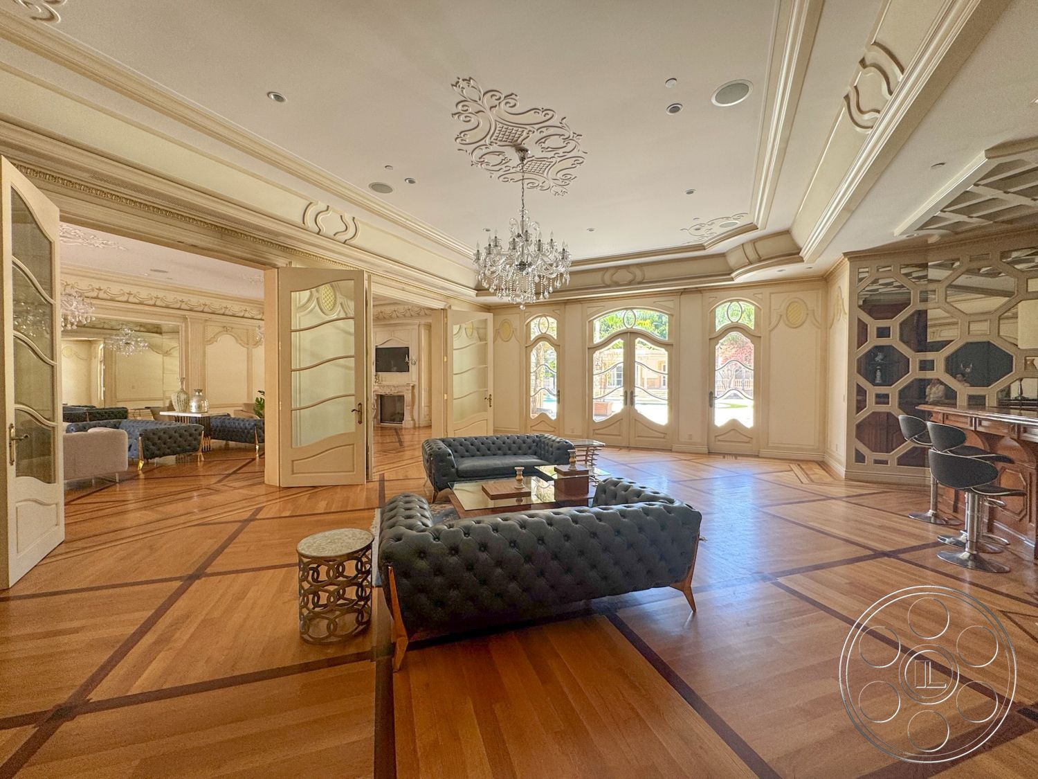 Tiffany Estate - chandelier lighting, open-concept living space, natural light from large windows, stylish coffee table, decorative molding details, glass double doors, decorative wall mirrors, modern bar area with stools, luxurious interior design, hardwood flooring with geometric inlay, ornate coffered ceiling, upholstered velvet sofas