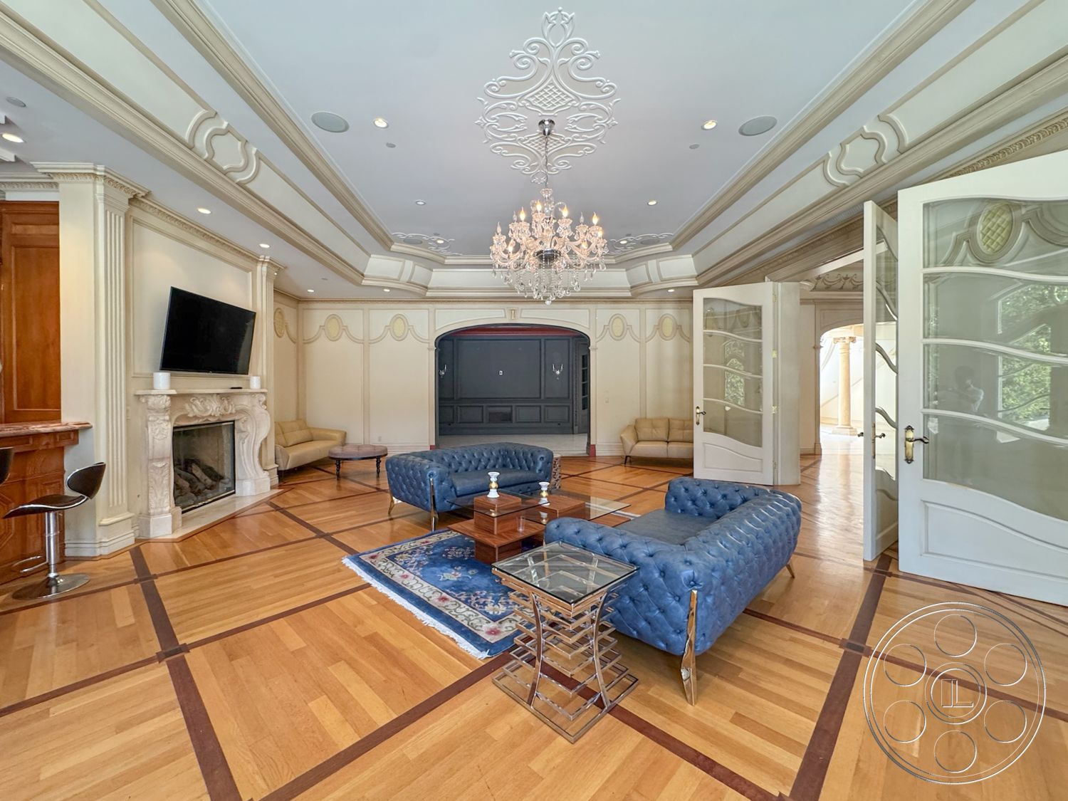 Tiffany Estate - chandelier lighting, decorative wall paneling, hardwood living room floors, marble fireplace surround, crown molding details, modern glass coffee table, decorative plaster ceiling with intricate details, double doors with glass panels, upholstered blue tufted sofas