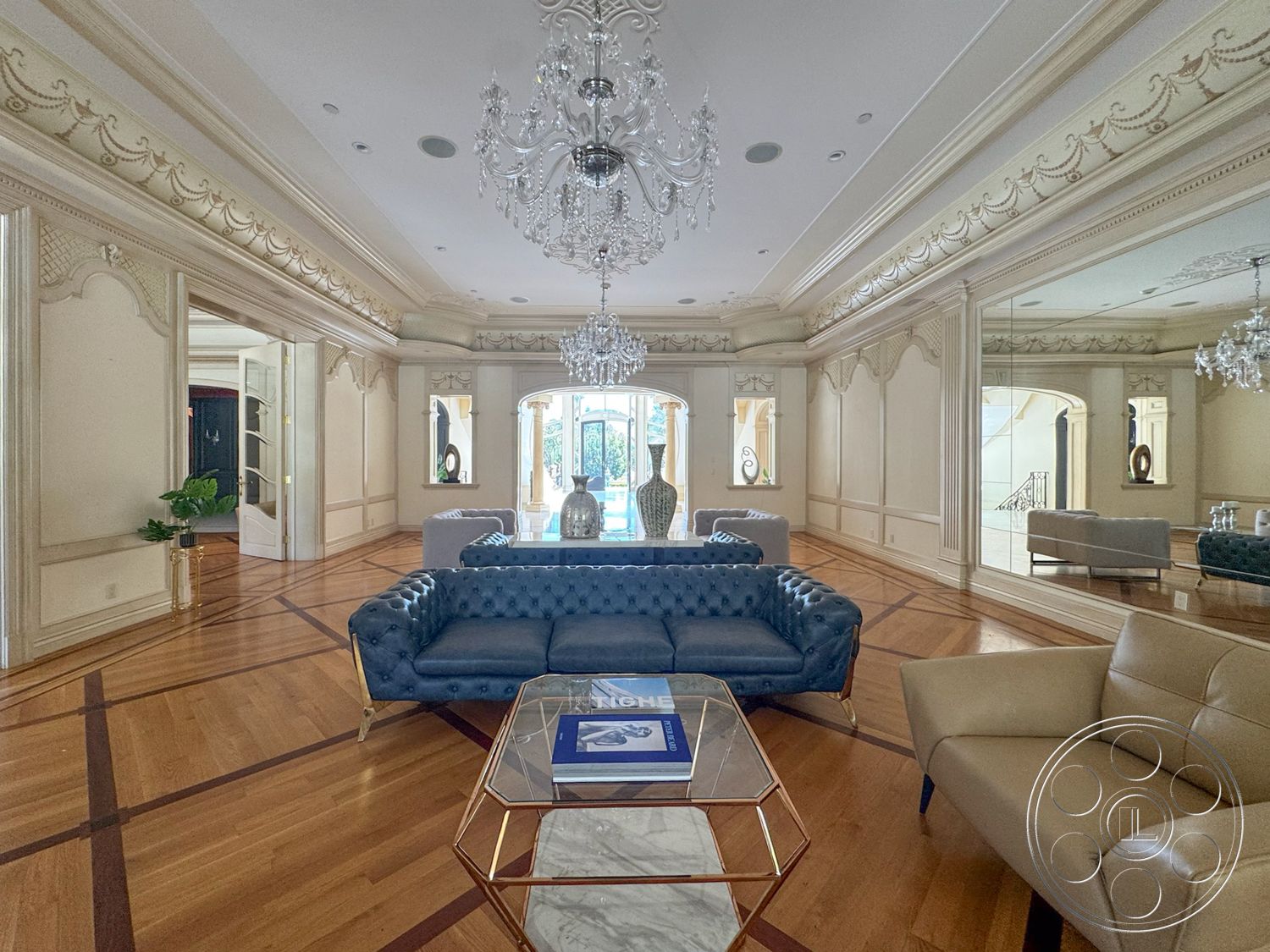 Tiffany Estate - hardwood flooring, spacious living area, natural light from large windows, artwork on display, decorative crown molding details, glass coffee table with marble top, ornate ceiling design, mirrored wall accents, open doorway leading to outdoor area, tufted blue velvet sofa, luxury chandelier, stylized white vase, plush beige armchair, intricate wall paneling