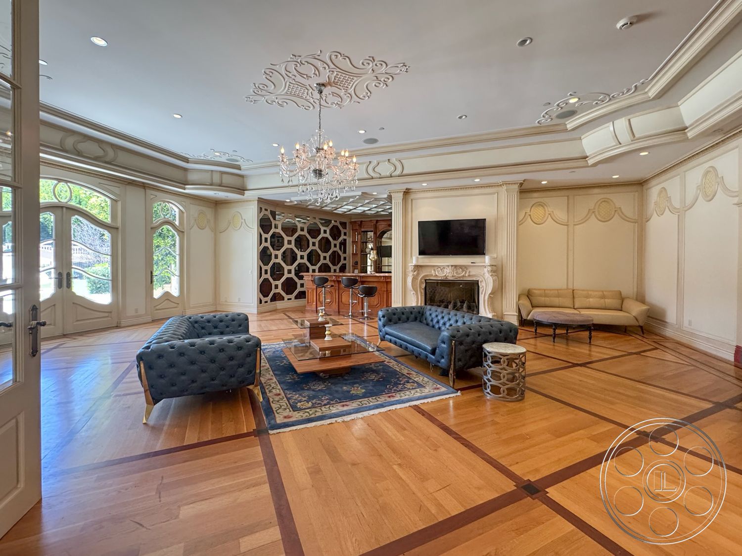 Tiffany Estate - hardwood flooring, decorative area rug, chandelier lighting, large arched windows, stone fireplace surround, ornate ceiling moldings, built-in bar area, tufted velvet sofa, custom wood feature wall, framed wall paneling, double paneled doors