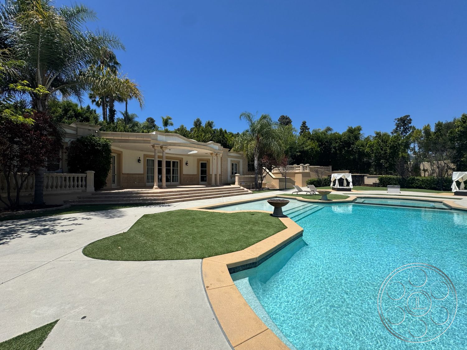 Tiffany Estate - exterior, paved driveway, palm trees, outdoor seating area, rectangular swimming pool, decorative stone wall, balustrade, stucco exterior walls, green lawn area, concrete pool border, concrete patio floor, modern outdoor design, lush landscaped garden, stone steps leading to pool area, under sunny blue sky