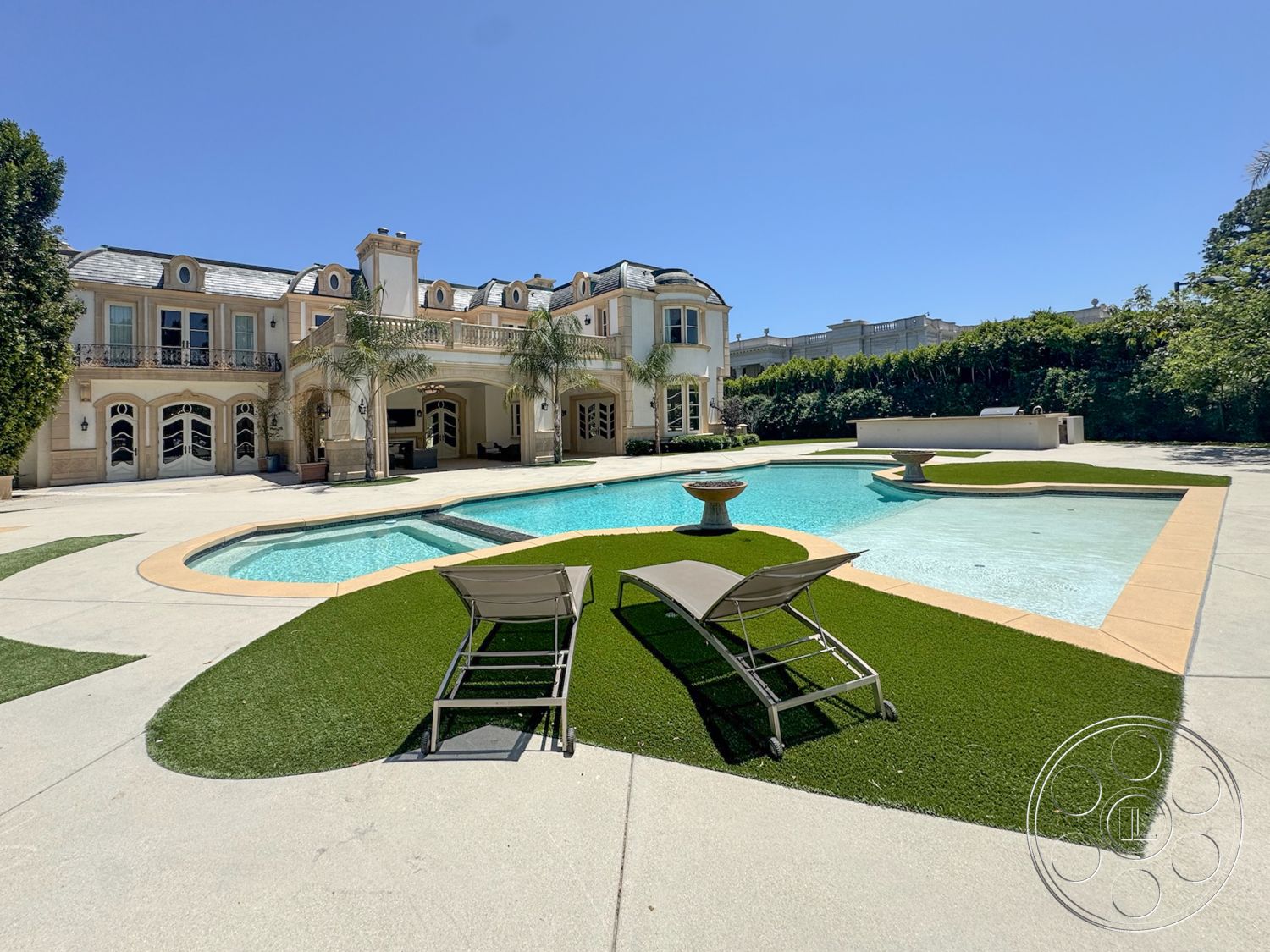Tiffany Estate - exterior, rectangular swimming pool, artificial grass landscaping, luxurious outdoor seating, sunny outdoor setting, concrete patio flooring, stucco exterior walls, arched window frames, textured concrete pool border, palm trees in landscape design, ornate balcony railings, plastered fountain feature, classic Mediterranean architecture