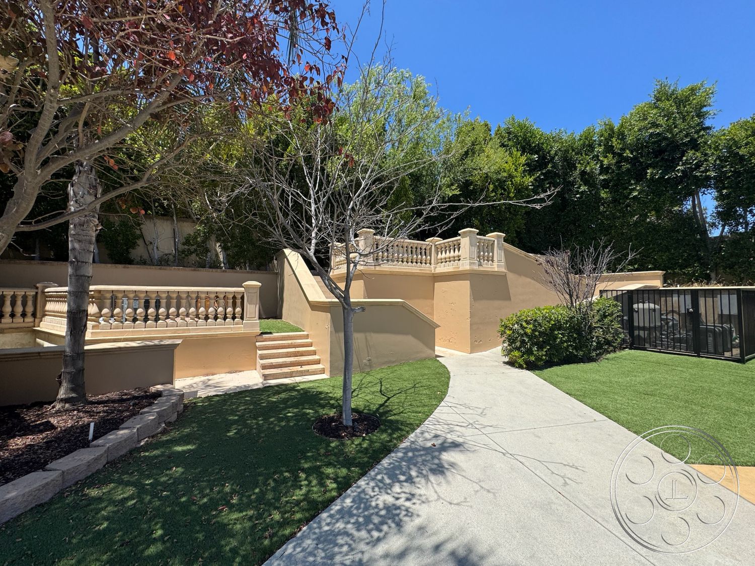 Tiffany Estate - exterior, lush green landscaping, stucco wall finish, decorative stone border, manicured shrubbery, artificial grass lawn, textured concrete walkway, ornate balustrade railing, rectangular pathway, granite concrete steps, tree-lined garden area
