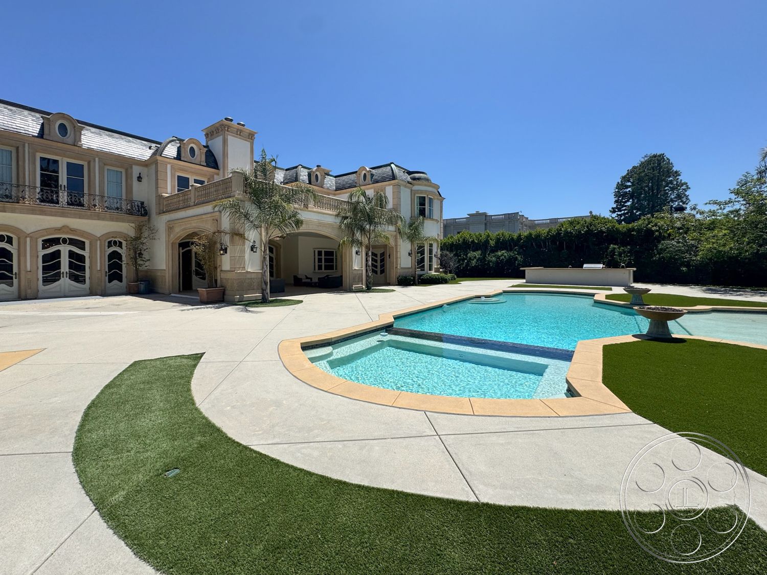 Tiffany Estate - exterior, landscaped garden, gabled roof, tropical palm trees, concrete pool deck, rectangular pool, stone fountain, modern residential architecture, paver walkway, stucco wall finish, synthetic grass lawn, interlocking stone patio flooring, ornate wrought iron balcony, masonry columns, expansive outdoor area, continuous blue tile pool border, elaborate outdoor structure