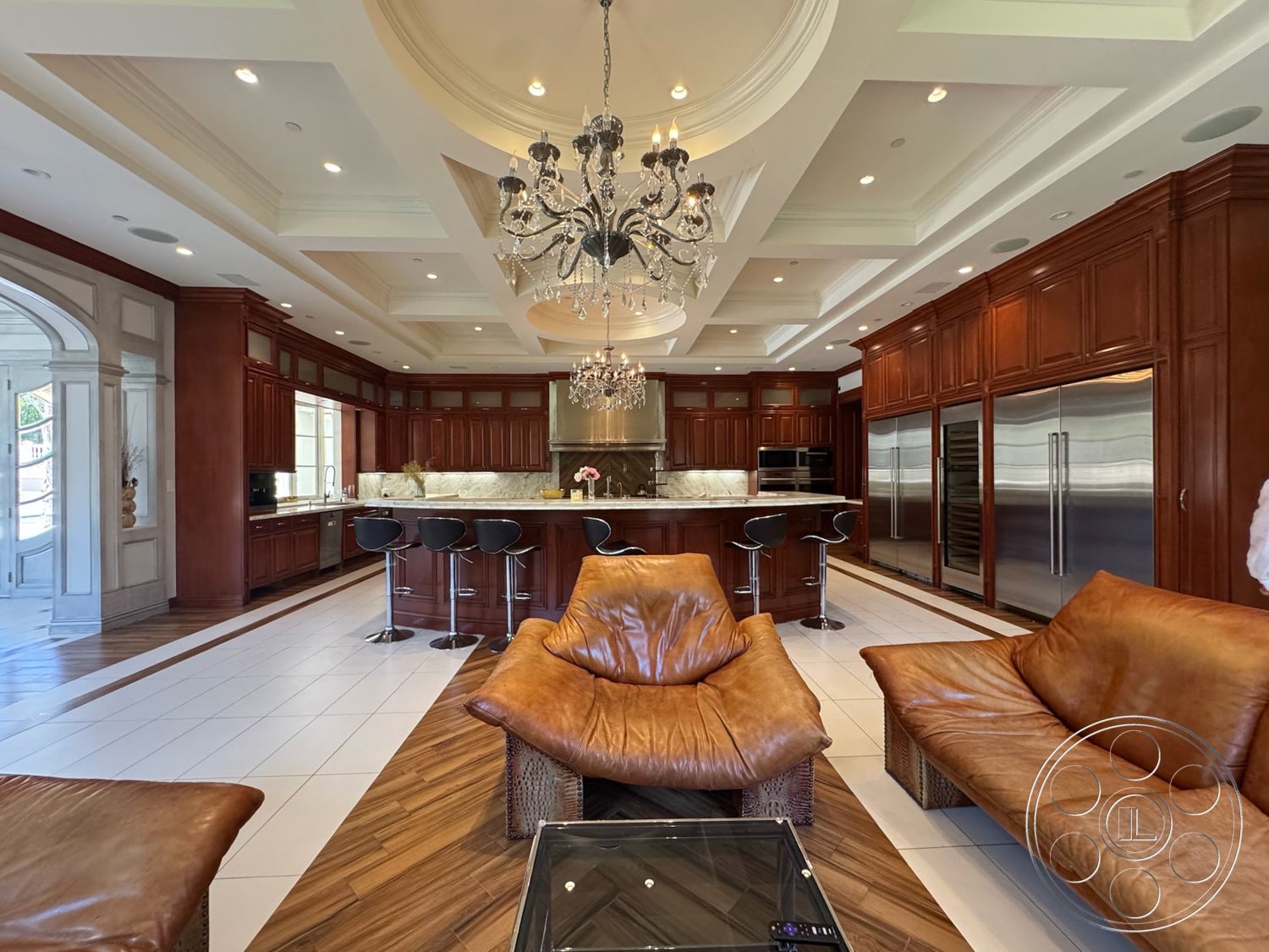Tiffany Estate - dark wood cabinetry, open concept design, black bar stools, dark wood dining table, brown leather sofas, chandelier light fixture, stainless steel kitchen appliances, light beige tile flooring, exposed coffered ceiling, white marble kitchen countertops, ornate door architrave, raised kitchen island, gleaming backsplash details