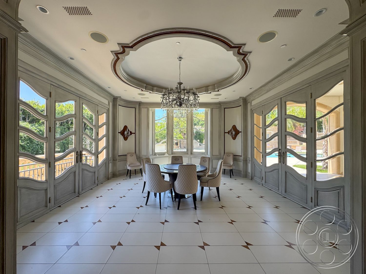 Tiffany Estate - bright natural light, round dining table, modern chandelier lighting, circular coffered ceiling, white porcelain tile flooring, geometric pattern tile detail, exposed ceiling with decorative molding, large windows with decorative frames, ornate double doors with wave glass panels, gray wood paneling on walls, upholstered dining chairs with light fabric, wall-mounted decorative mirror, view overlooking landscaped outdoor space