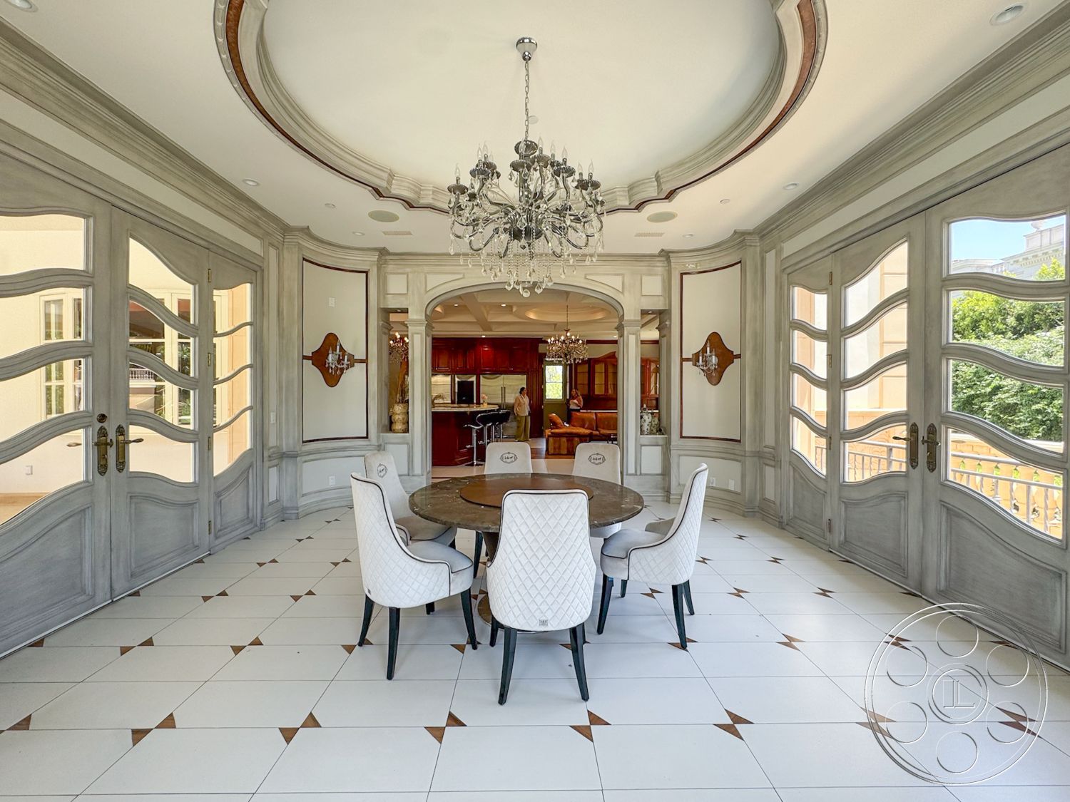 Tiffany Estate - natural light from large windows, open-concept layout, decorative wall panels, chandelier lighting fixture, rounded coffered ceiling, cabinets in cherry wood finish, white tile flooring with geometric accents, glass double doors with ornate details, round dining table in dark wood, upholstered dining chairs with quilted pattern, elaborate wall moldings, luxurious dining area