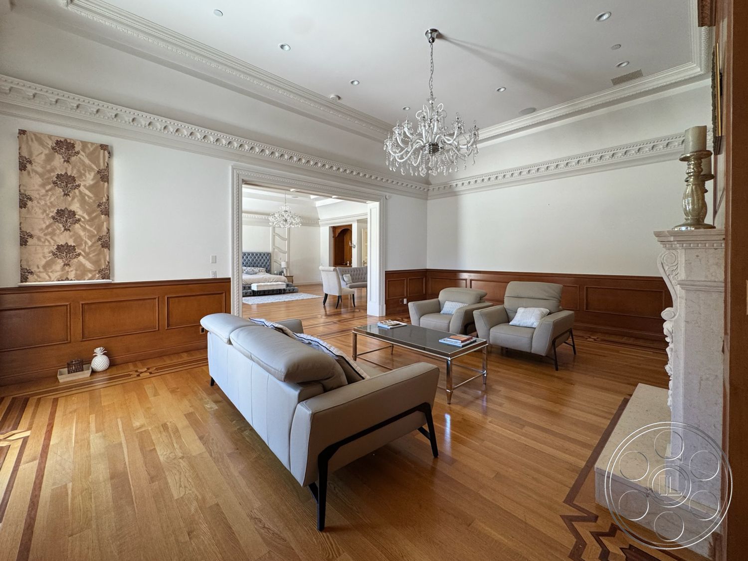 Tiffany Estate - hardwood flooring, open floor plan, glass coffee table, chandelier lighting, natural light from windows, dark wood paneling, luxurious living room, decorative wall hanging, white painted walls, marble fireplace surround, crown molding details, contemporary seating arrangement, elegant room design