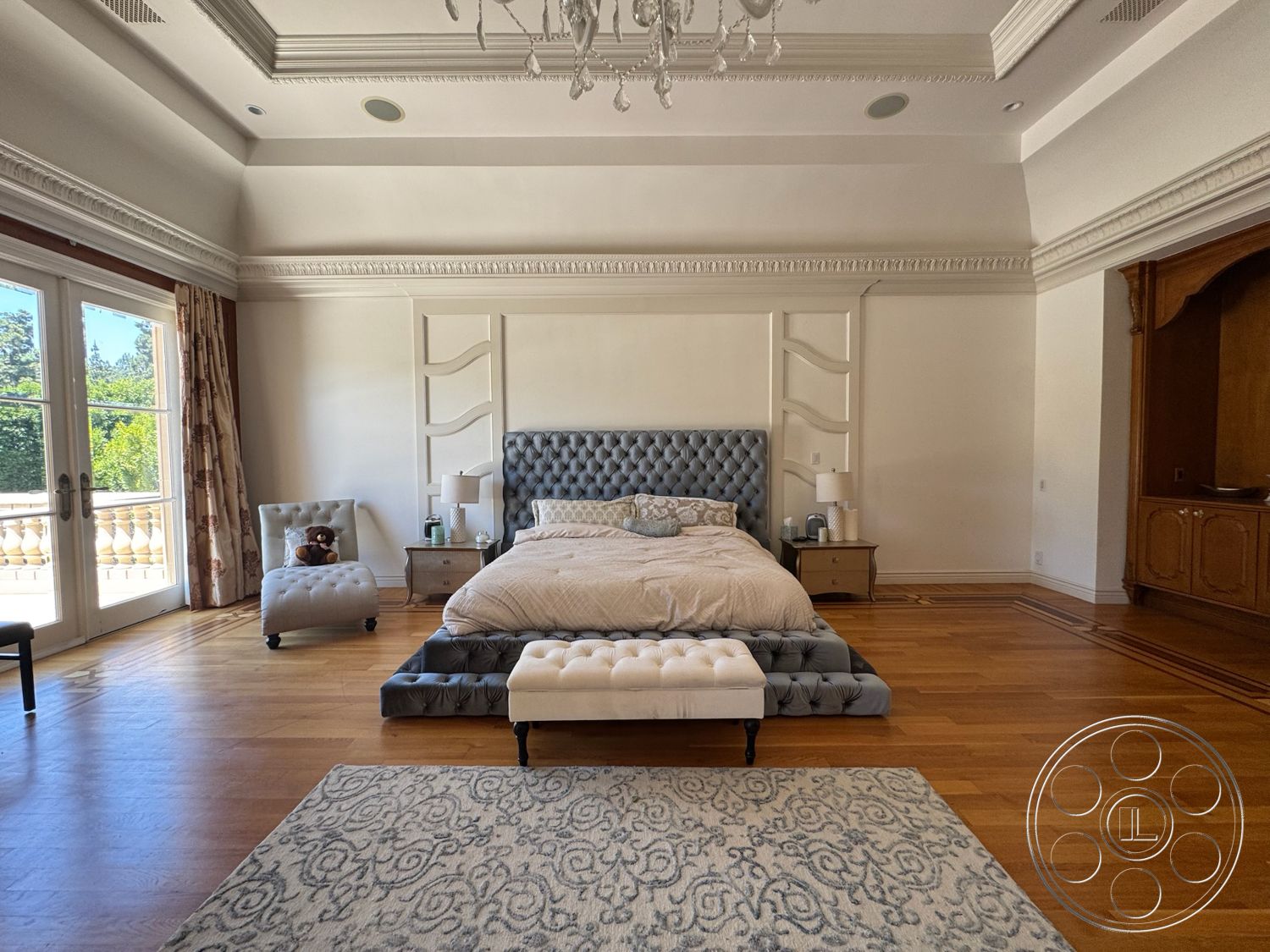 Tiffany Estate - open floor plan layout, hardwood bedroom floors, decorative area rug with intricate pattern, white painted walls, built-in wooden cabinetry, luxurious master bedroom, coffered ceiling with crown molding details, tufted upholstered bed with gray fabric headboard, drapery with floral pattern, glass French doors leading to outdoor balcony, side tables with lamps