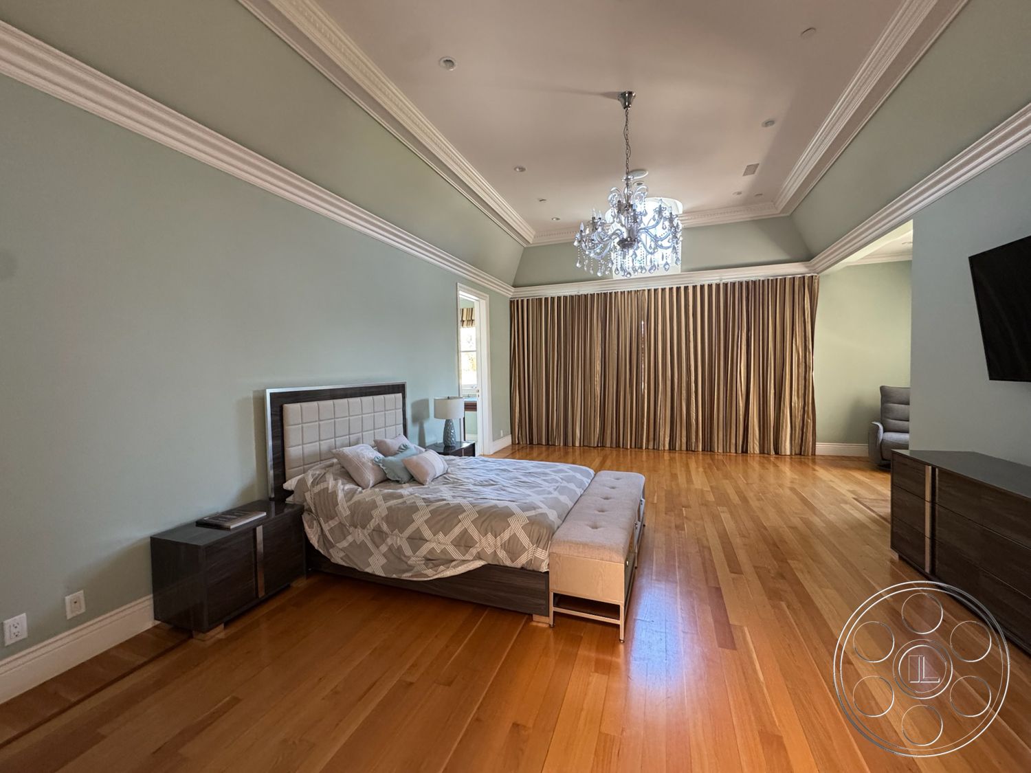 Tiffany Estate - master bedroom, chandelier light fixture, hardwood bedroom floors, sitting area with modern chair, modern bed with upholstered headboard, contemporary television unit, coffered ceiling with crown molding, pale green painted walls, elegant floor-to-ceiling drapes, dark wood nightstands