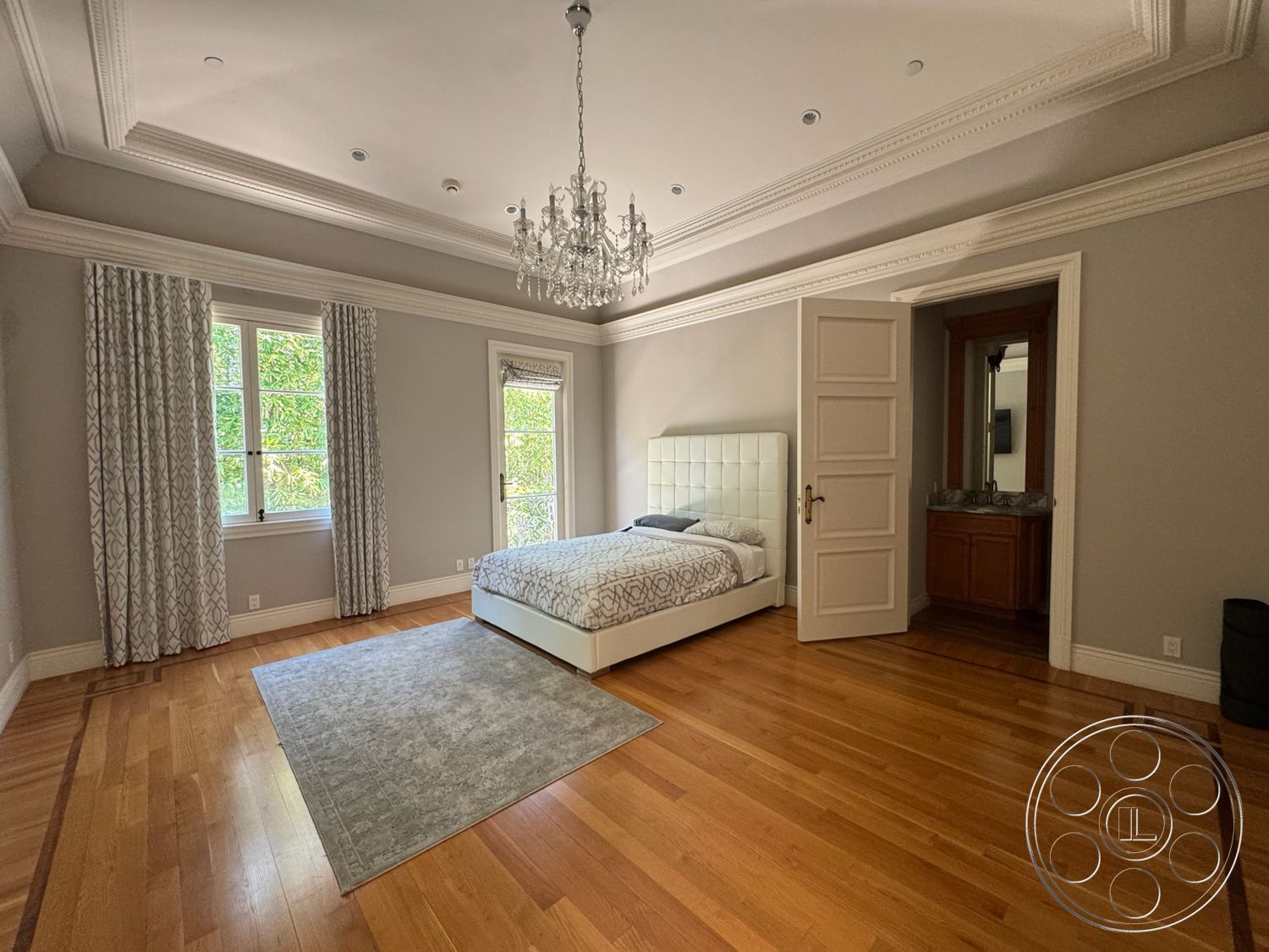 Tiffany Estate - natural light, coffered ceiling, floor-to-ceiling windows, beige area rug, hardwood bedroom floors, crown molding details, wooden bedroom door, light grey painted walls, king-size upholstered bed, gilded chandelier, door with detailed woodwork, grey patterned curtains, vanity with wooden cabinets, bedroom setting