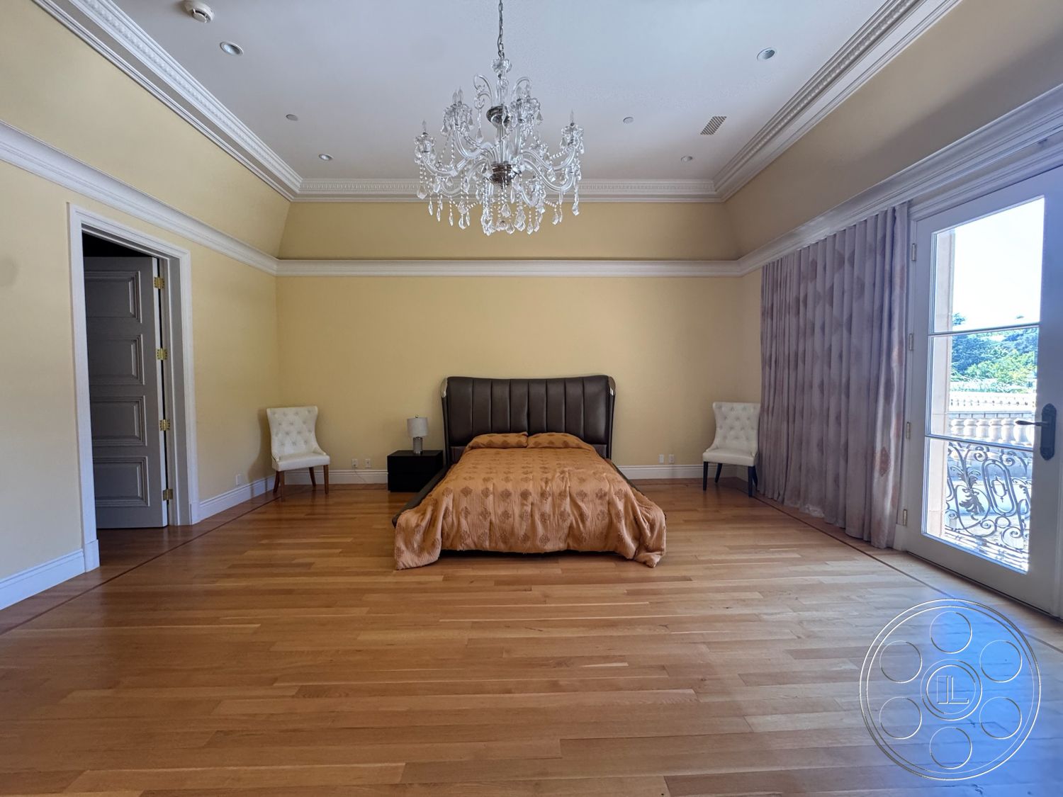 Tiffany Estate - floor-to-ceiling windows, vaulted ceiling, chandelier lighting, French doors, hardwood bedroom floors, light yellow painted walls, crown molding details, sheer curtain drapes, tan upholstered bed, decorative accent chair