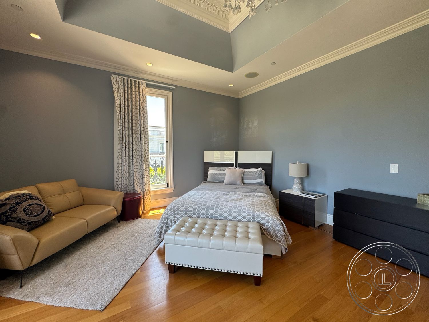 Tiffany Estate - decorative area rug, modern lamp, recessed ceiling lighting, hardwood bedroom floors, gray painted walls, modern leather sofa, white crown molding, contemporary bedside table, sheer window curtains, upholstered bed with geometric pattern bedding, tufted white ottoman, glass window with urban view, minimalist dresser