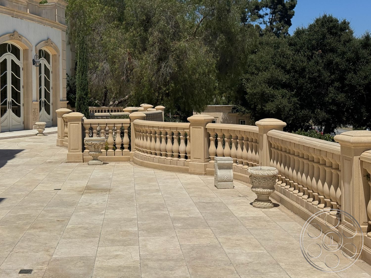 Tiffany Estate - exterior, surrounding trees, textured stone walls, lush green landscaping, sunlit outdoor space, potted plant accent, travertine patio flooring, ornate stone balustrade, arched entry doors, elegant outdoor terrace