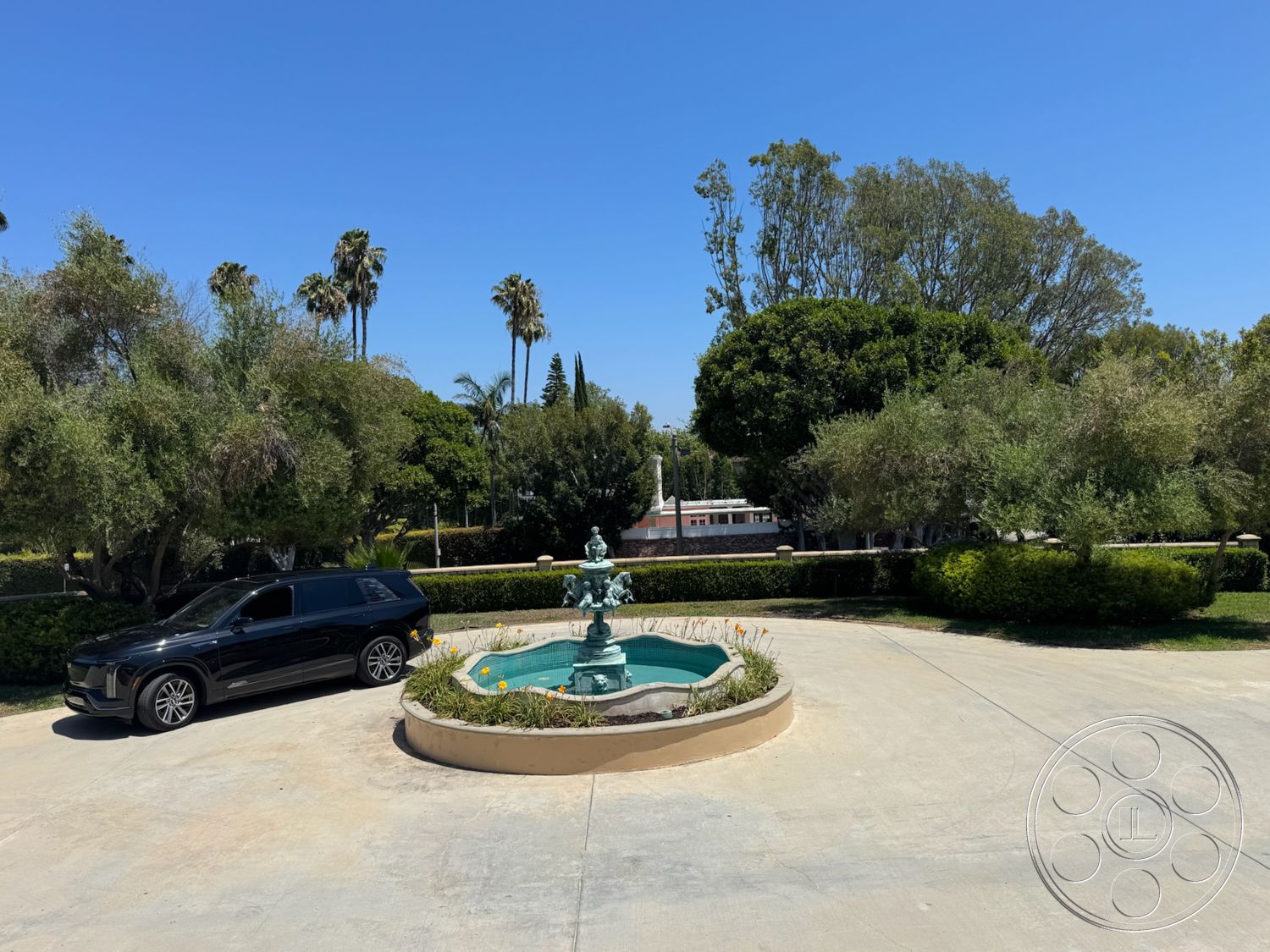 Tiffany Estate - exterior, palm trees, concrete driveway, flowering plants, landscaped garden beds, sunny suburban environment, circular fountain with turquoise water, fountain with bronze detailing, lush green bushes, blue sky view
