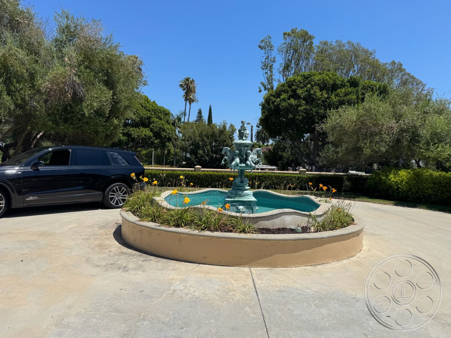 Tiffany Estate - exterior, lush greenery, large trees, concrete driveway, tall palm trees, yellow flowering plants, landscape planting, rounded flower bed, sculptural fountain, stone fountain basin, dramatic blue sky