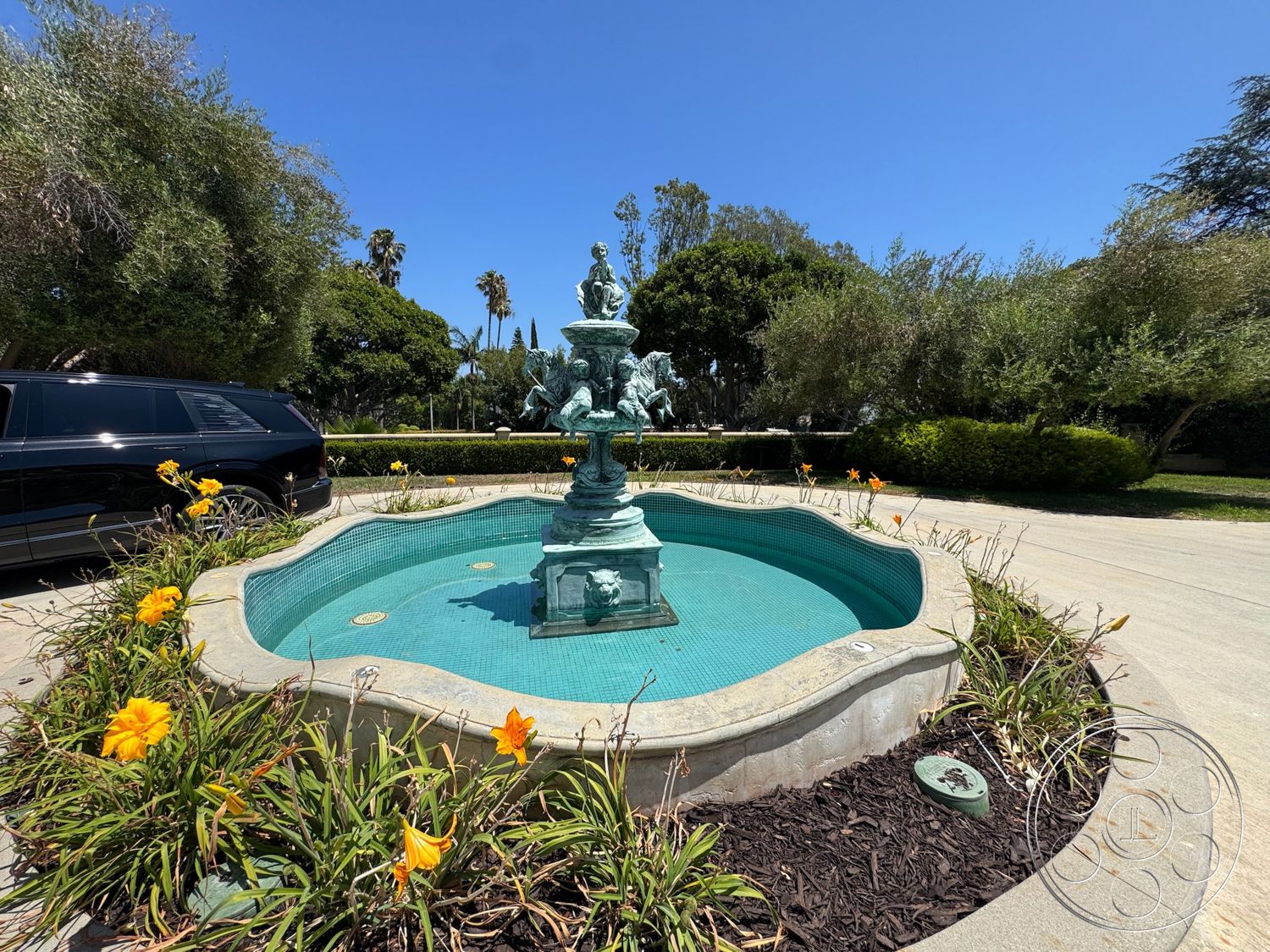 Tiffany Estate - exterior, concrete driveway, paved stone path, yellow flowering plants, landscaped flower beds, lush greenery surroundings, tile fountain basin, bronze fountain sculpture, curved stone pool border, detailed water feature, ornamental garden design