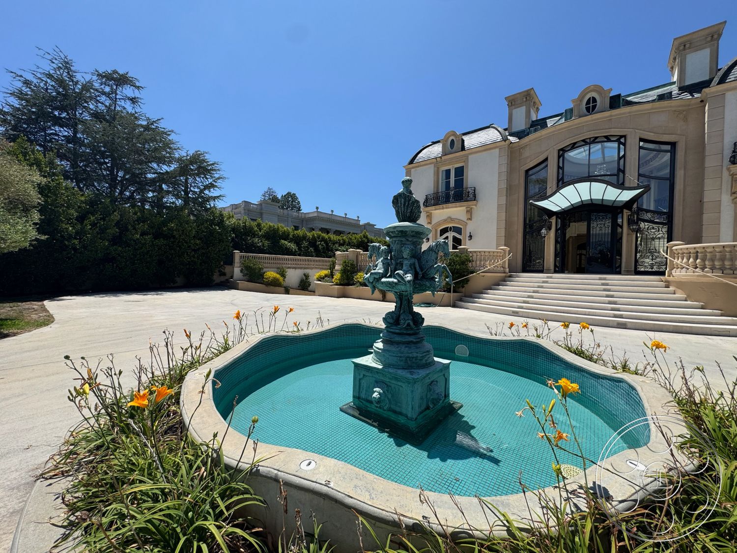 Tiffany Estate - exterior, landscaped greenery, large floor-to-ceiling windows, smooth concrete driveway, stone patio flooring, symmetrical architectural design, ornate fountain with turquoise mosaic tiles, circular fountain shape, landscaped garden with yellow flowers, two-story Mediterranean-style home, arched entrance with glass awning, balcony railings with intricate metalwork, exposed stone accent walls