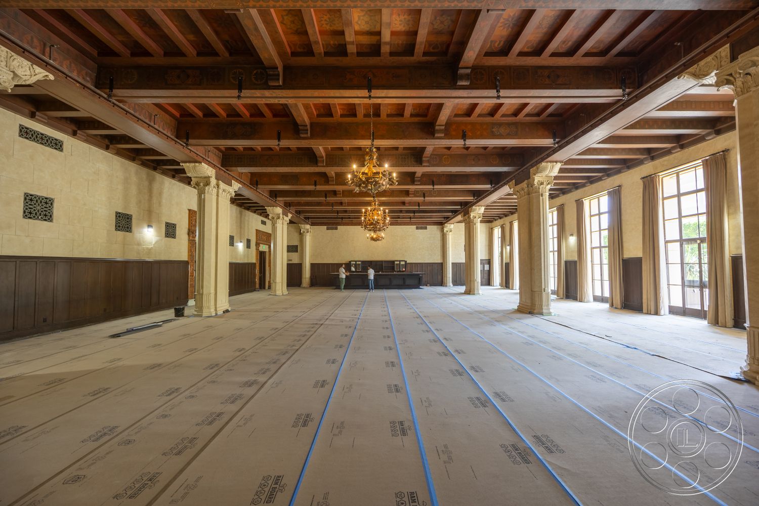 The Plaza - dark wood paneling, stone wall accents, exposed wood ceiling beams, coffered ceiling details, textured plaster wall treatments, open space interior, chandeliers with intricate designs, ornate column supports, large windows with classical frames, unfurnished ballroom setting