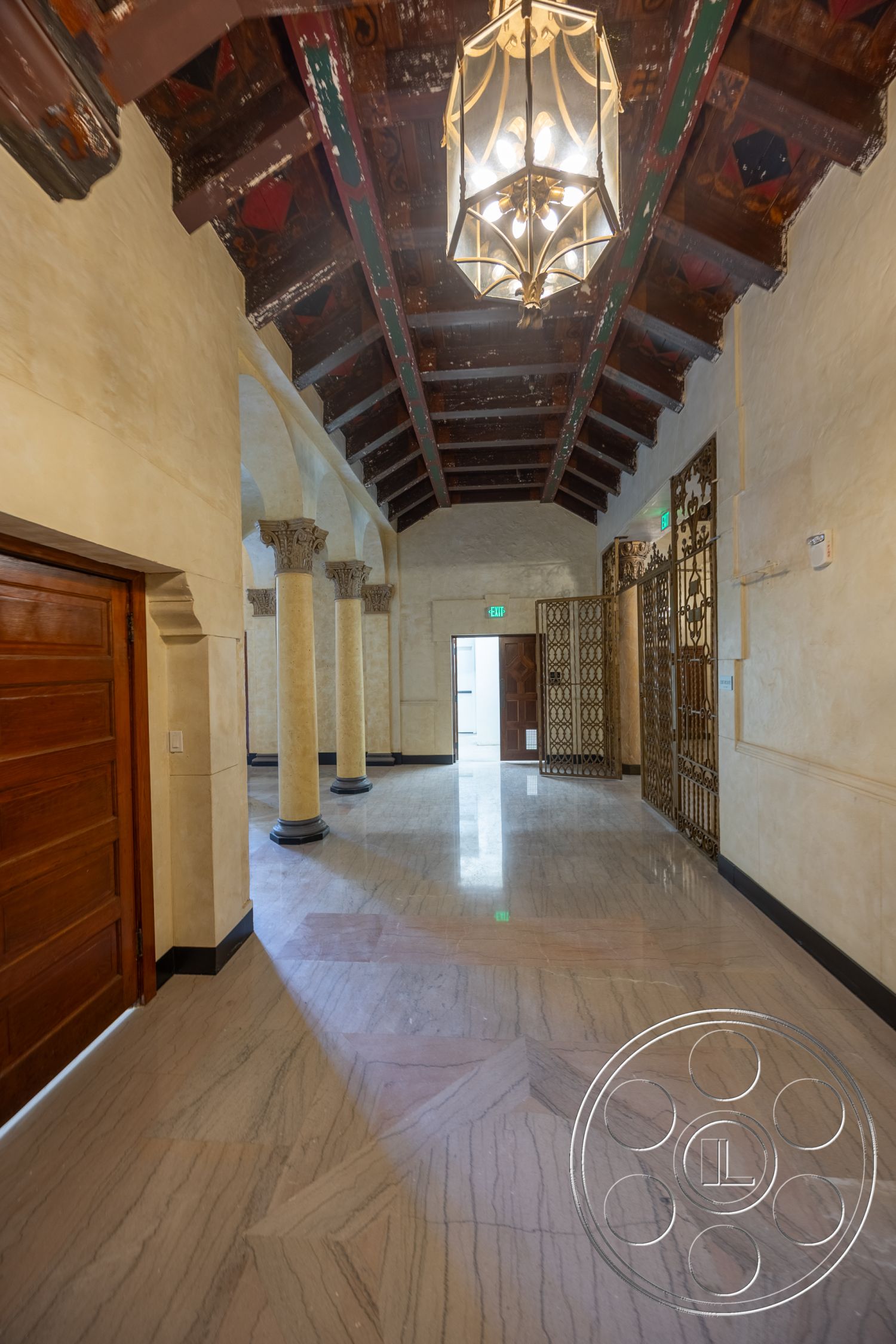 The Plaza - neutral color palette, marble flooring, arched doorways, textured plaster walls, traditional architectural style, wooden entry door, exposed wooden beam ceiling, ornate chandelier lighting, tall decorative columns, ornate metal gate