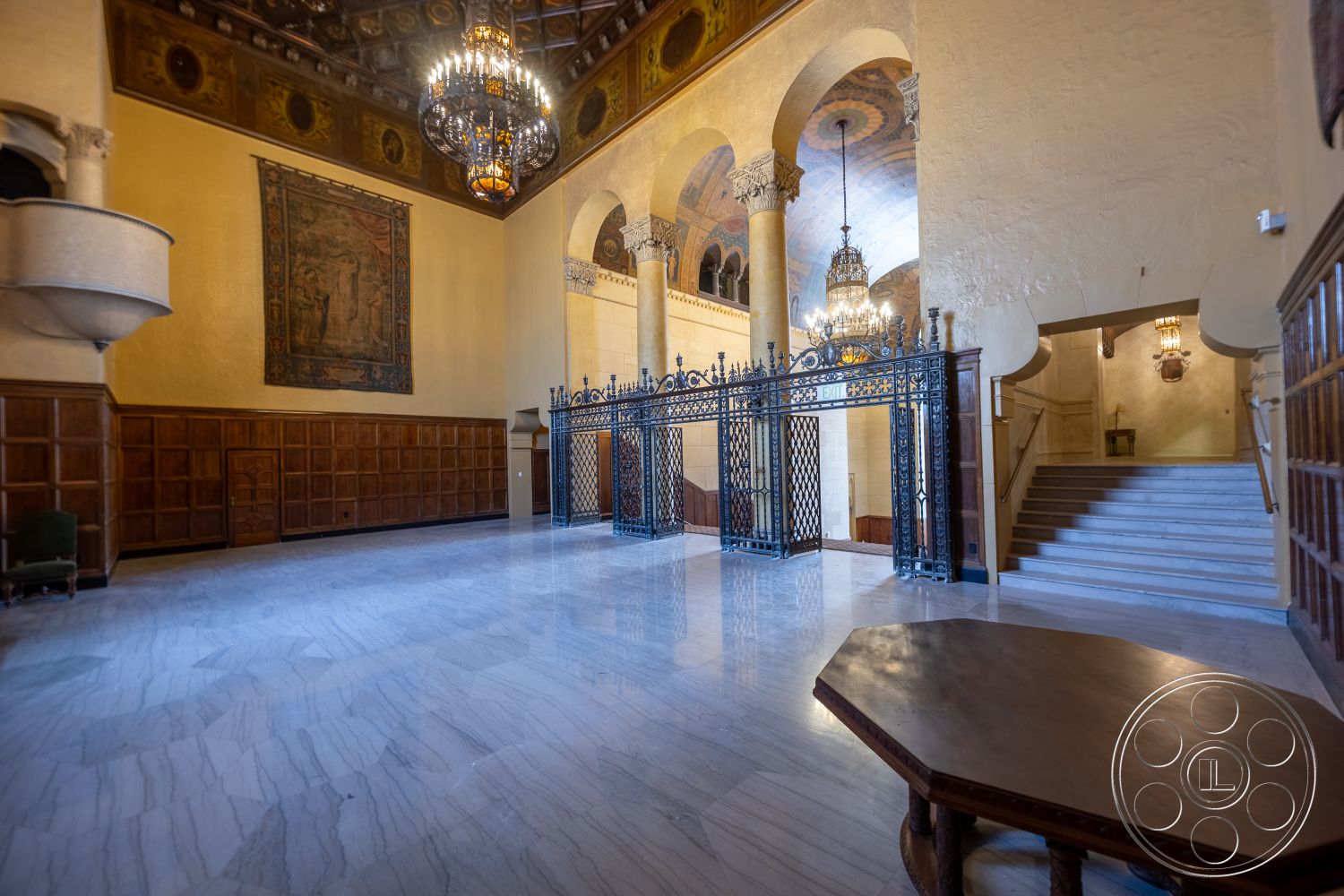 The Plaza - high ceilings, exposed wooden beams, vaulted ceiling, marble flooring, arched doorways, wood paneling walls, metal security gate, historic architectural style, ornate chandelier lighting, tapestry wall hanging, staircase with marble steps, dark wood accent details, decorative plaster details