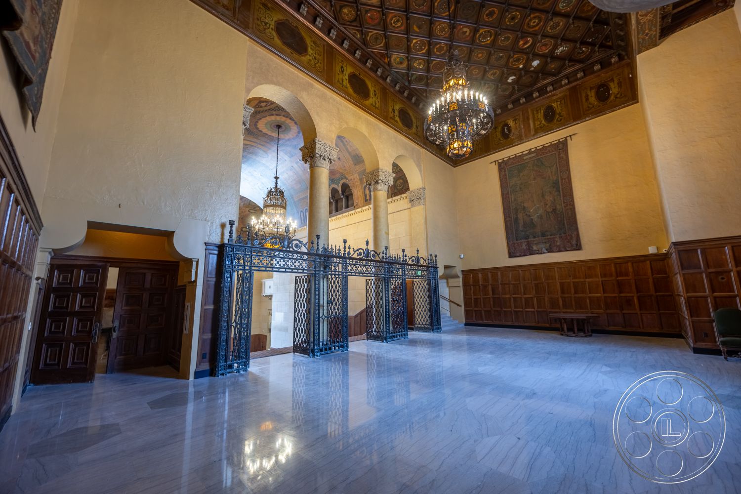 The Plaza - wood paneling, iron gate, vaulted ceiling, chandelier lighting, marble flooring, arched doorways, textured plaster walls, ornate ceiling details, historic architectural style, exposed columns, decorative tapestries, open foyer space