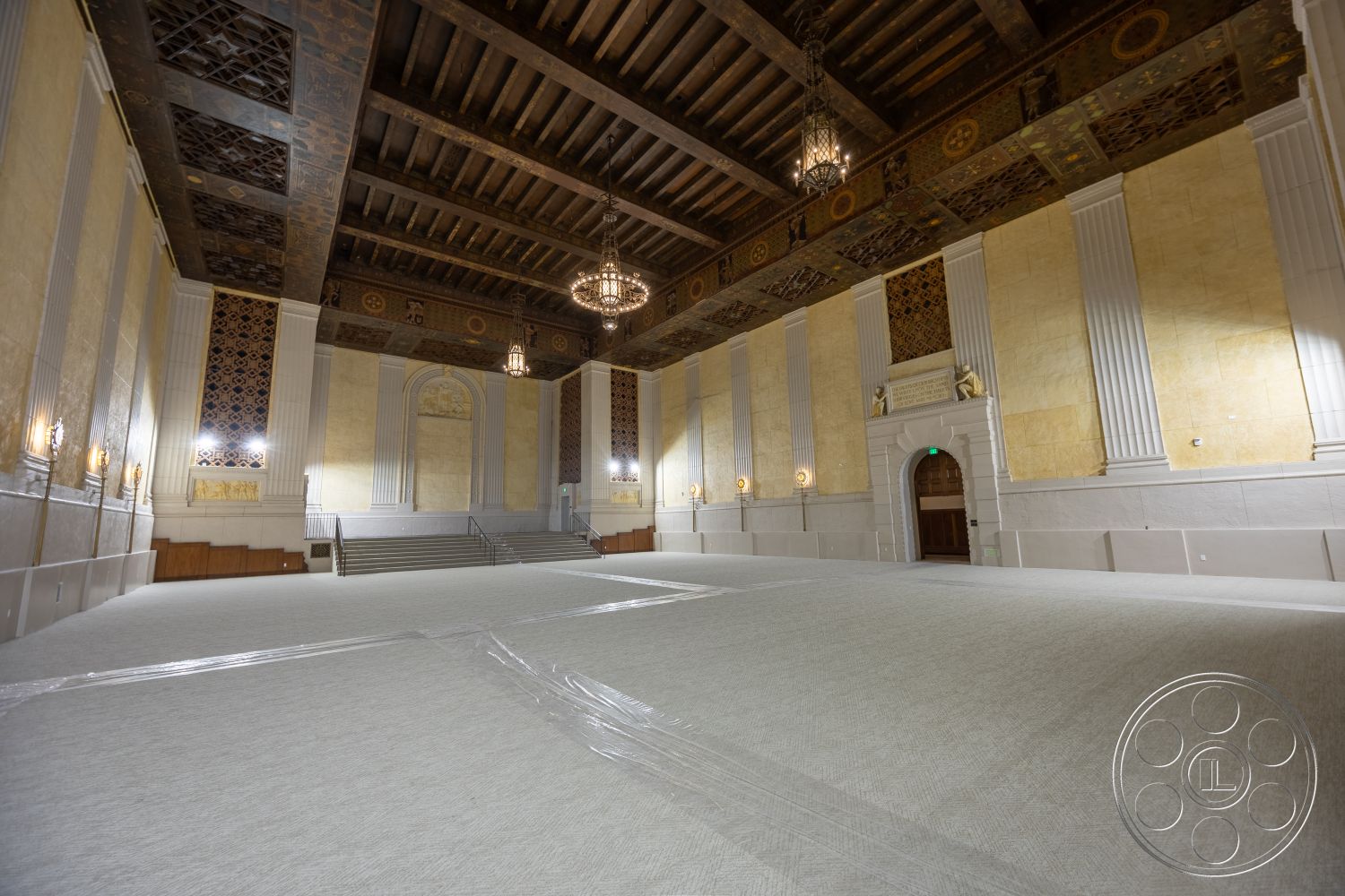 The Plaza - polished stone flooring, textured plaster walls, high vaulted ceilings, exposed wooden ceiling beams, wood paneling accents, neutral carpet flooring, decorative wall moldings, ornate plaster ceiling details, elegant wall sconces, gold leaf ceiling panels, lattice wood wall panels, chandeliers with vintage design, columned wall accents, arched doorways with decorative trim, built-in step seating area, large open floor space, grand entryway arch